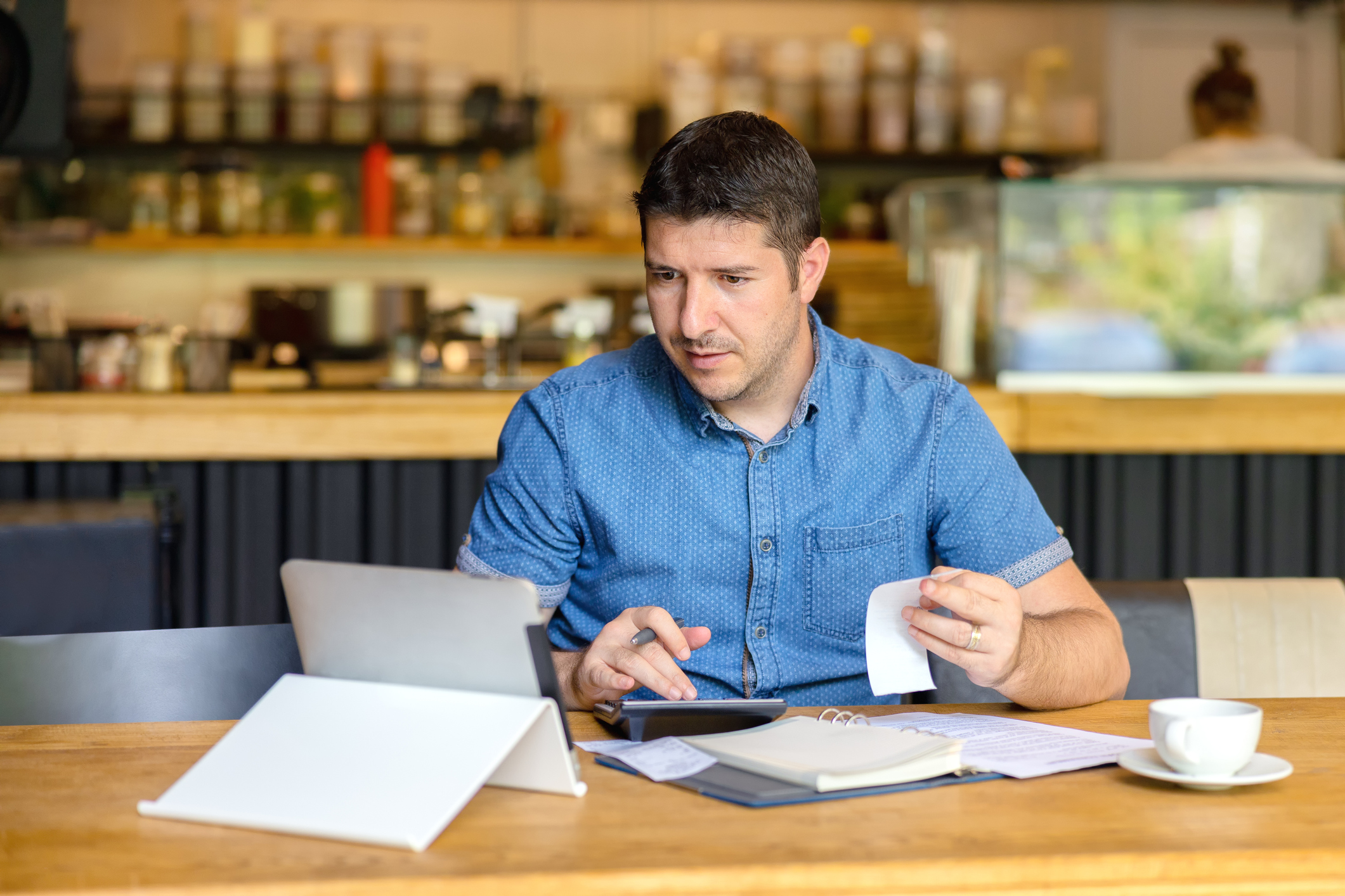 Man at table holding slip of paper and typing on calculator with laptop, notebook, and mug in front of him