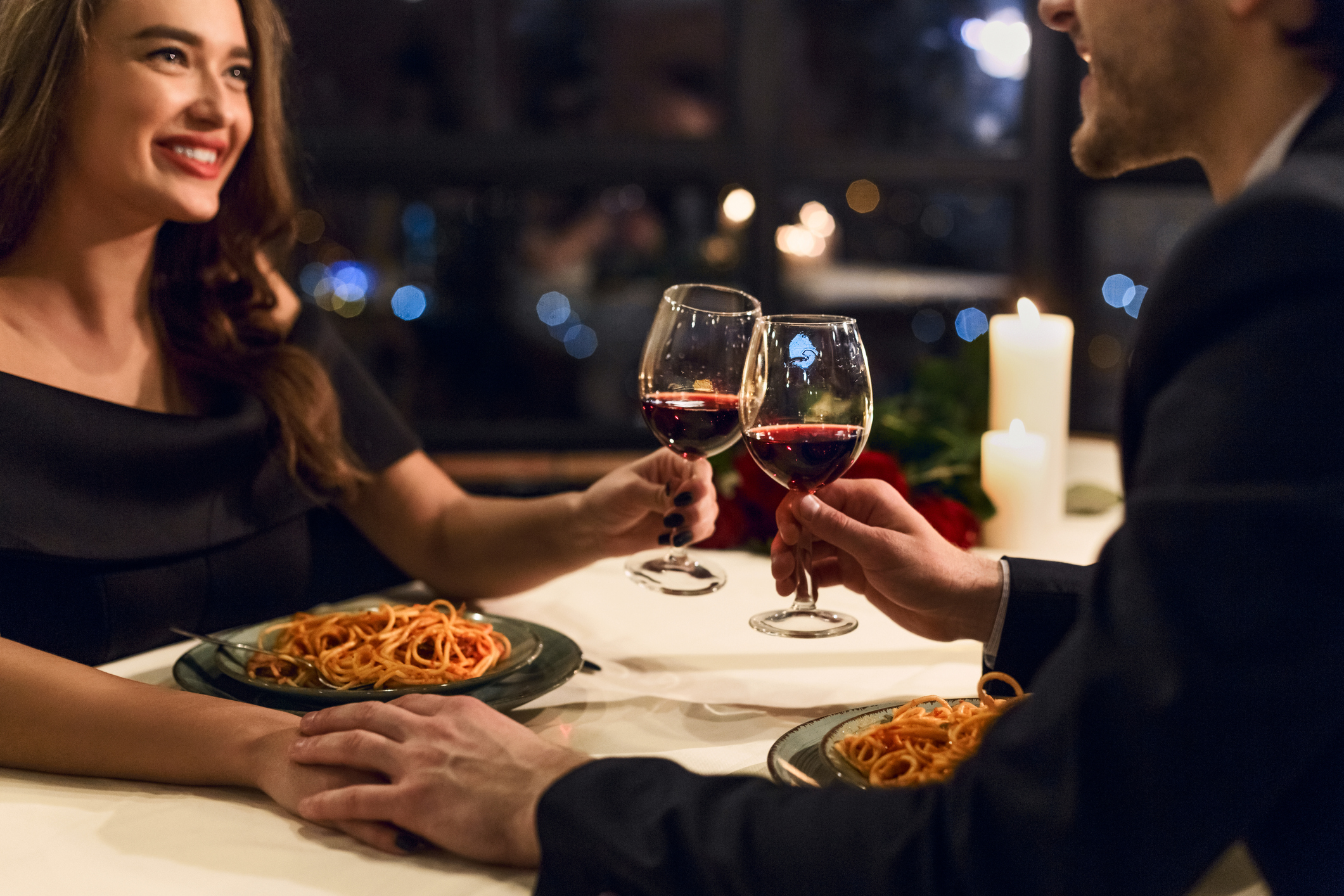 A man and a woman clink glasses of red wine and hold hands across a table, with plates of pasta in front of them