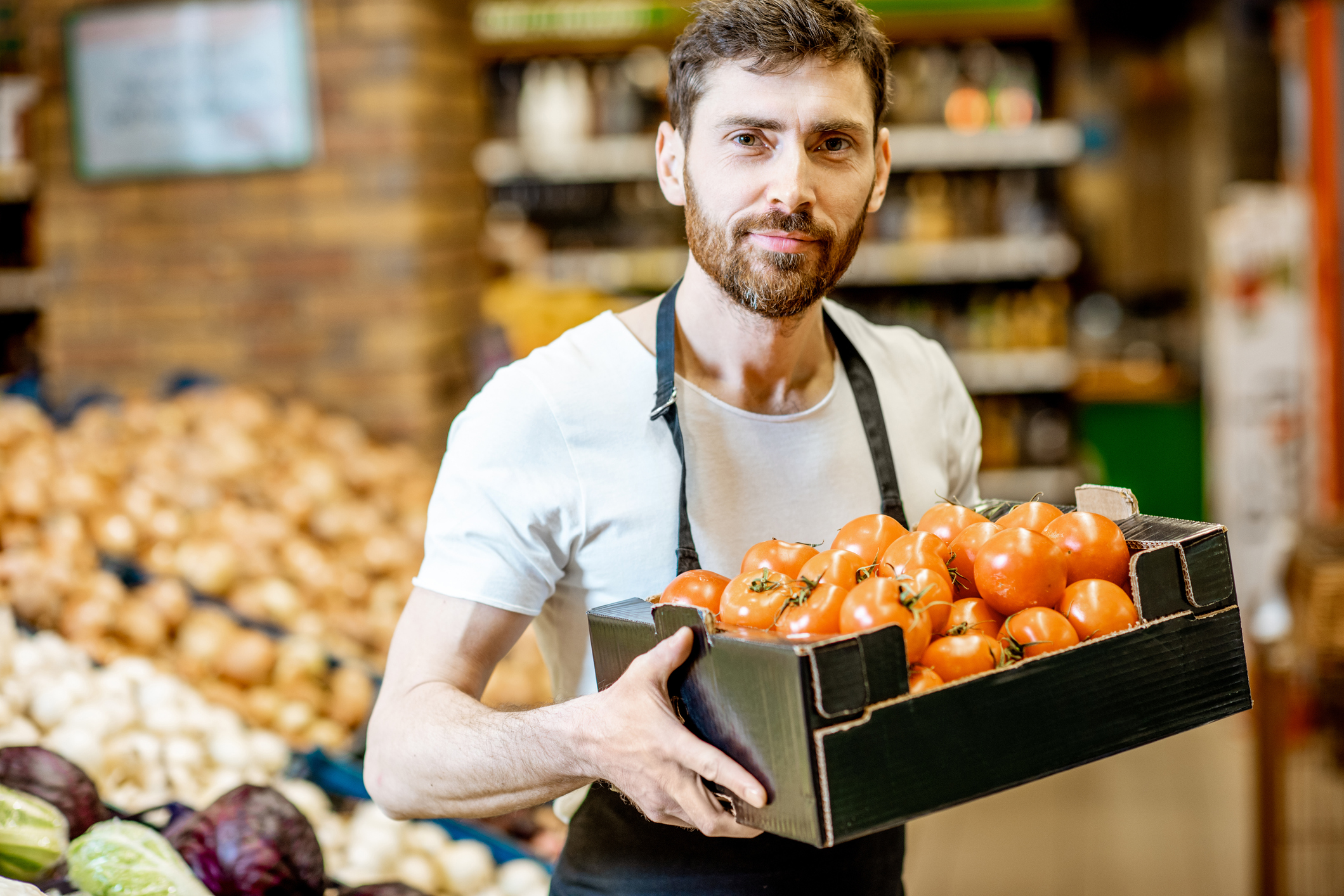 Man wearing apron in supermarket holding box of tomatoes