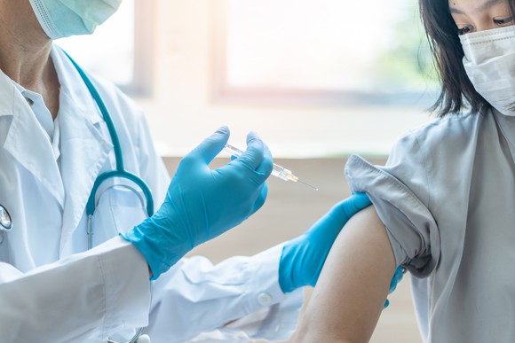 A person wearing a stethoscope and mask vaccinates a masked patient or volunteer.