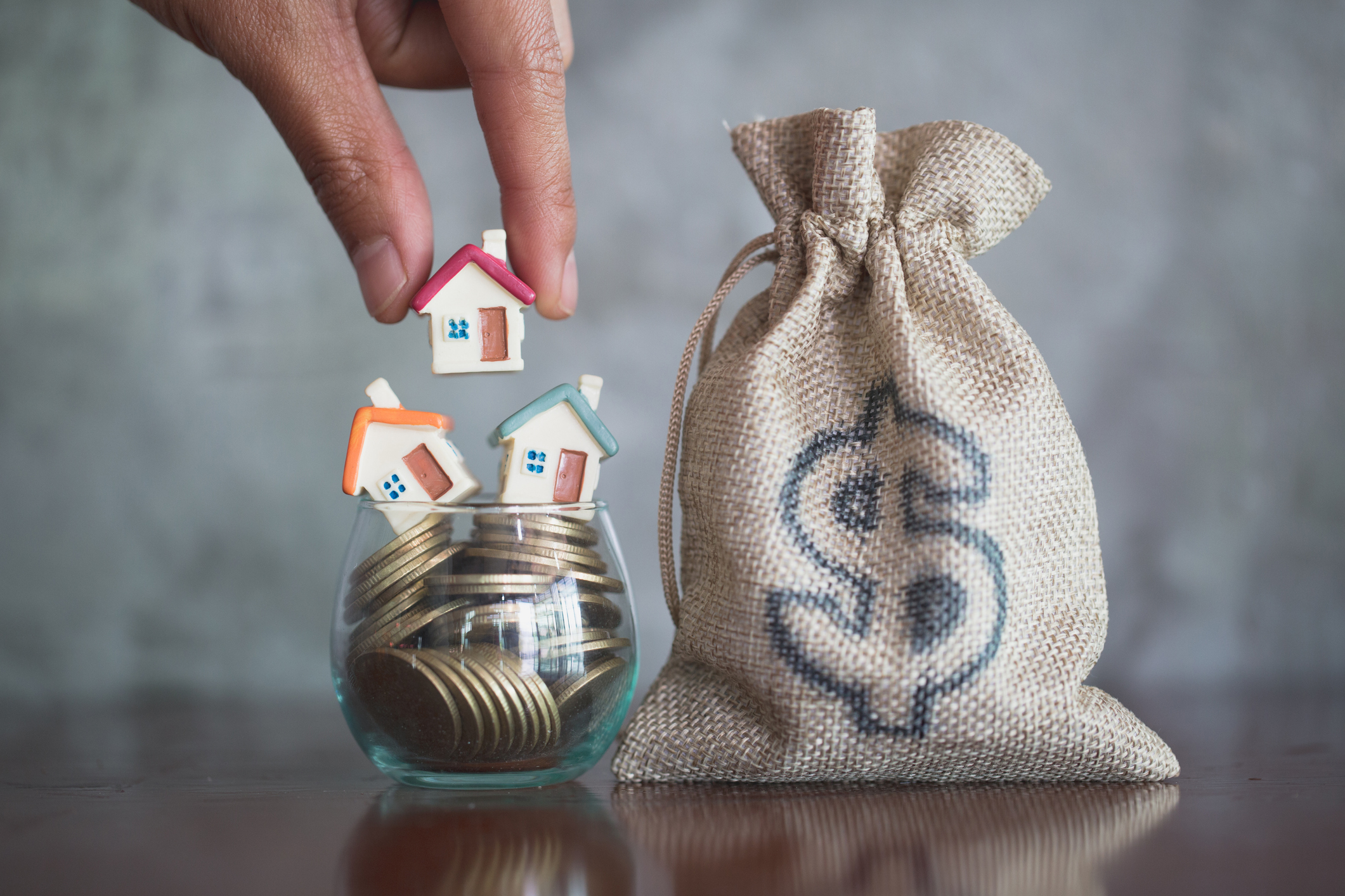 A hand placing miniature houses into a glass of coins next to a burlap money bag.
