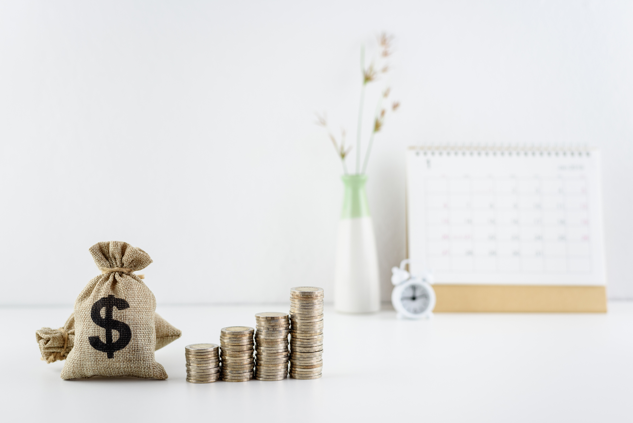 Bag of money with increasing stacks of coins with a calendar and clock in the background.