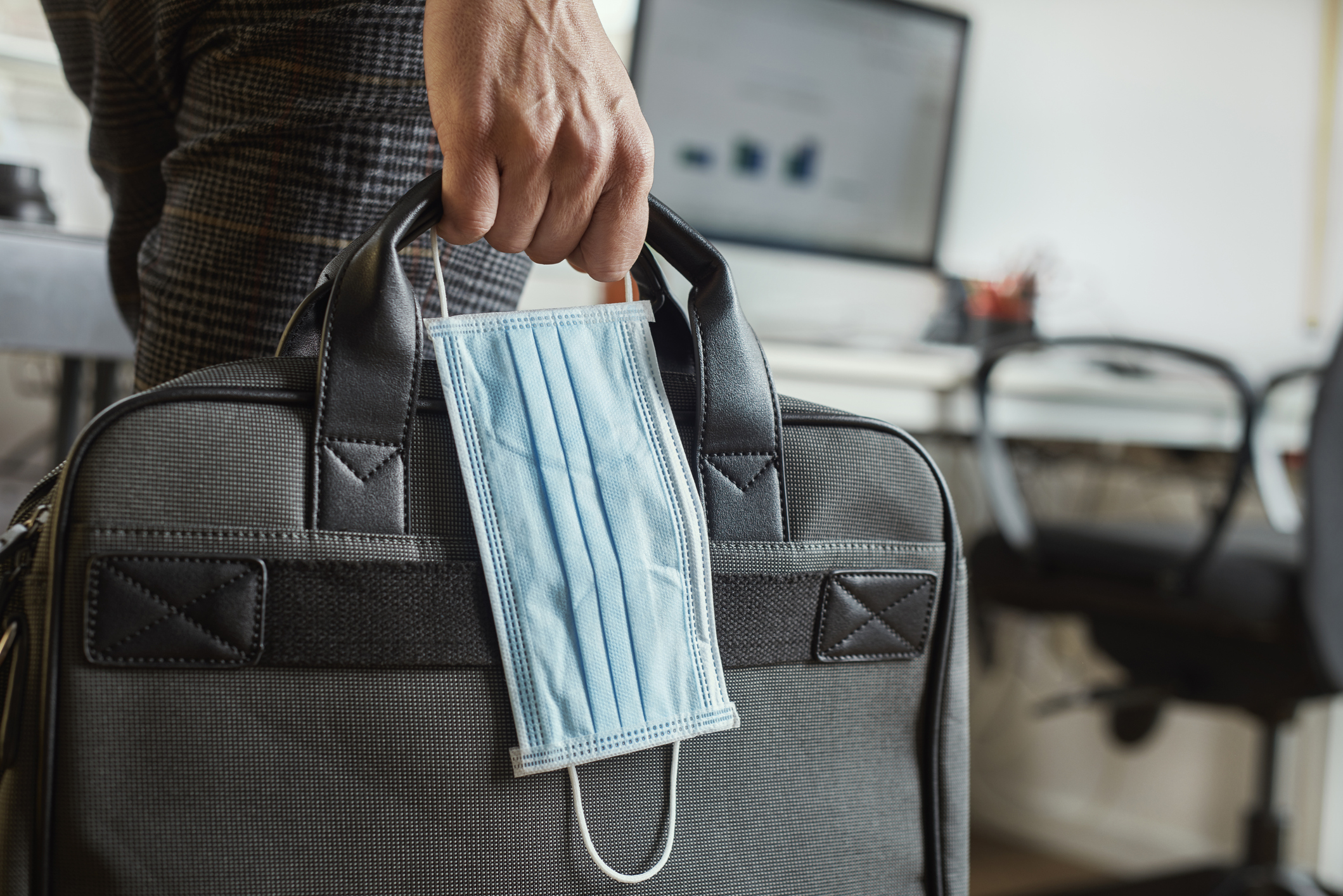 Closeup of a male hand holding a briefcase with a mask dangling from it.
