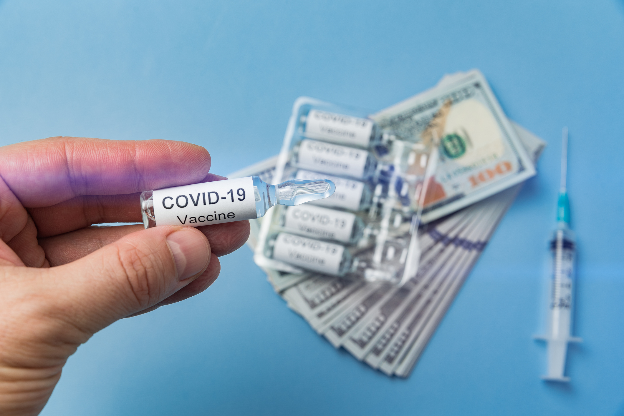 A hand holds a vial labeled COVID-19 vaccine with hundred dollar bills in the background.