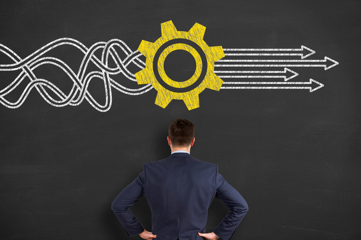 A businessman thinking while looking at a chalkboard with a gear converting tangled lines into straight arrows drawn on it.