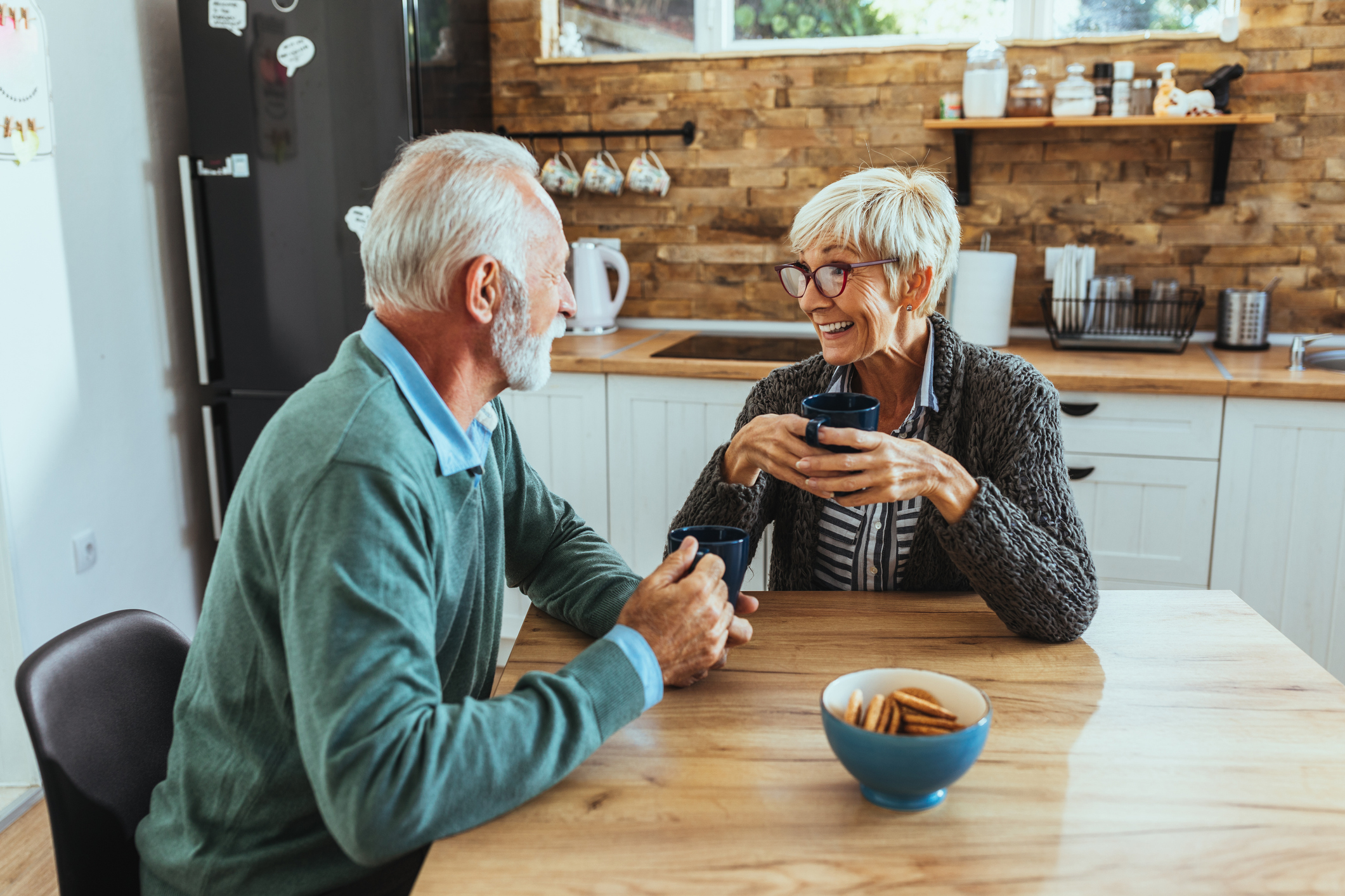 Older couple sitting at kitchen table holding mugs.
