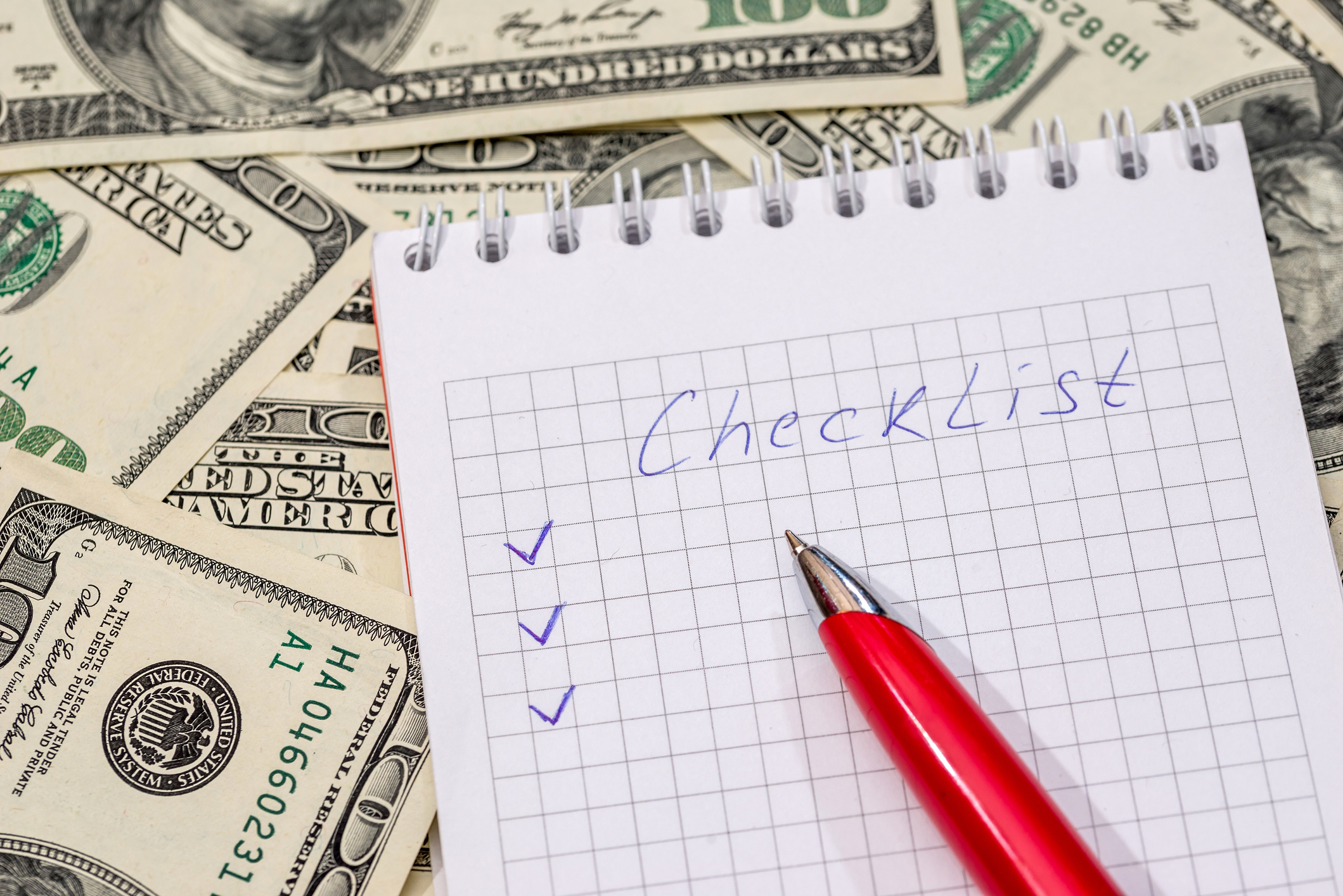 A pen sits on a notebook with the word "checklist" written on it. The notebook sits atop a pile of money.