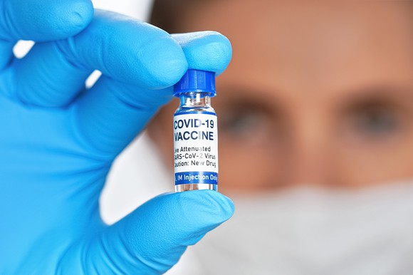 Masked person wearing disposable gloves and holding a vaccine vial labeled COVID-19 VACCINE