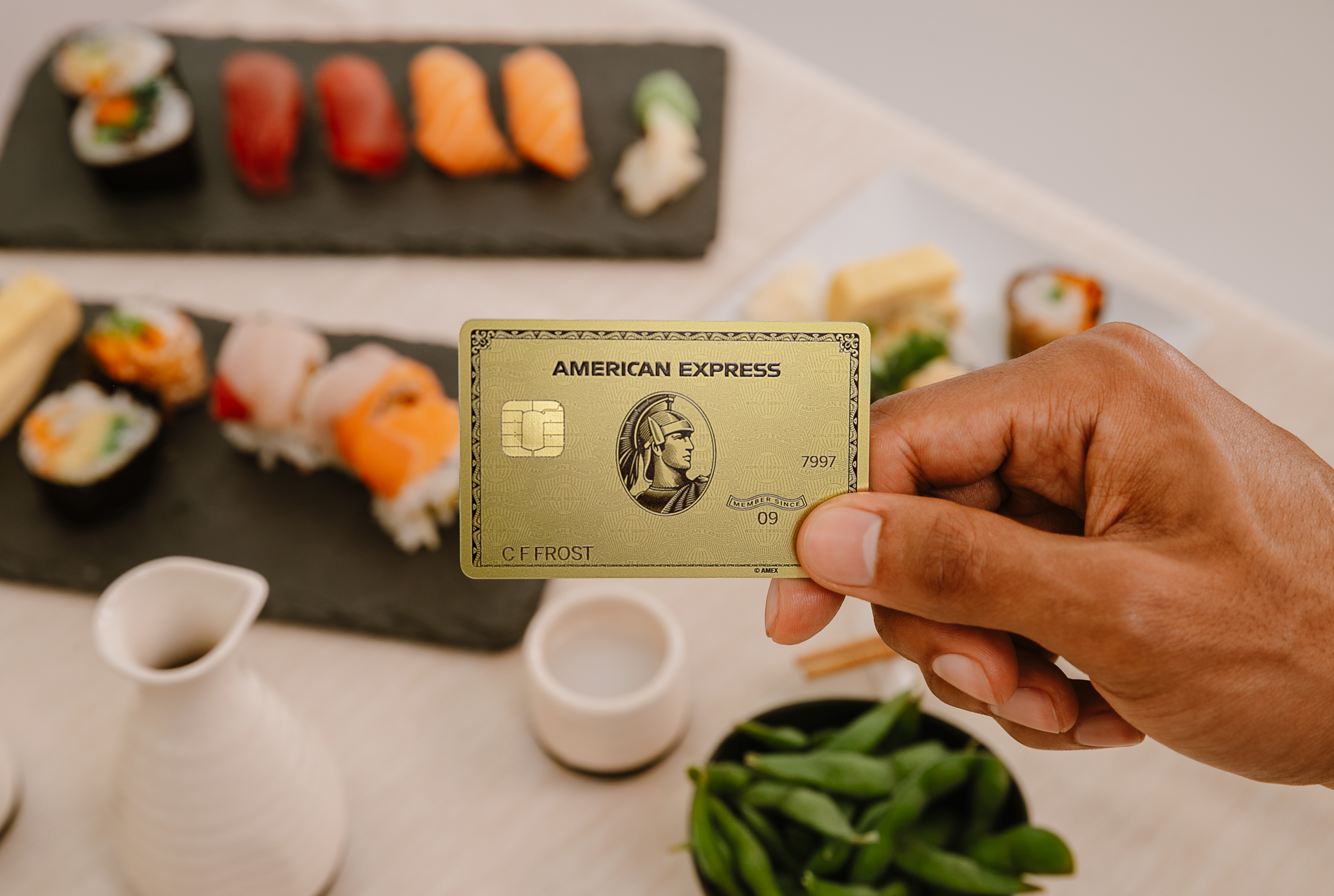 A hand holds an American Express gold card above a table full of food.