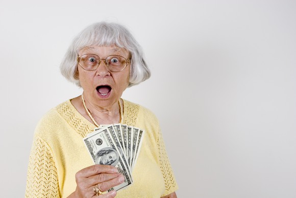 An older woman is holding a bunch of hundred dollar bills, with her mouth open in surprise.