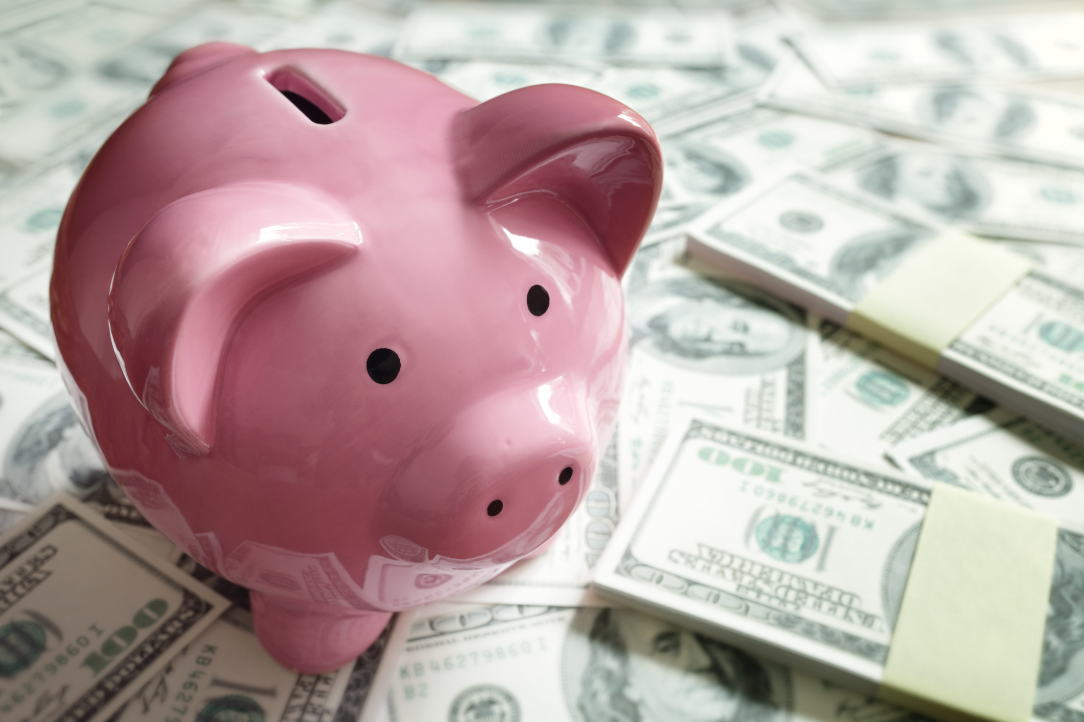 A piggy bank on top of hundred dollar bills spread around.