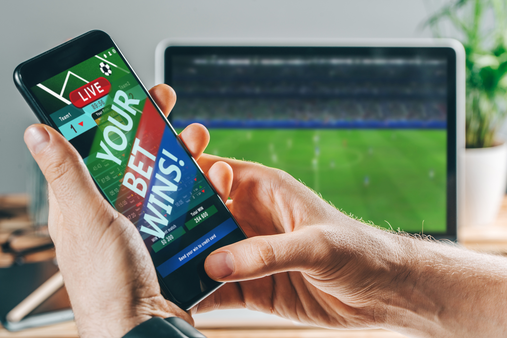 A phone with the words "your bet wins" on it and a soccer match playing on a screen behind the phone.