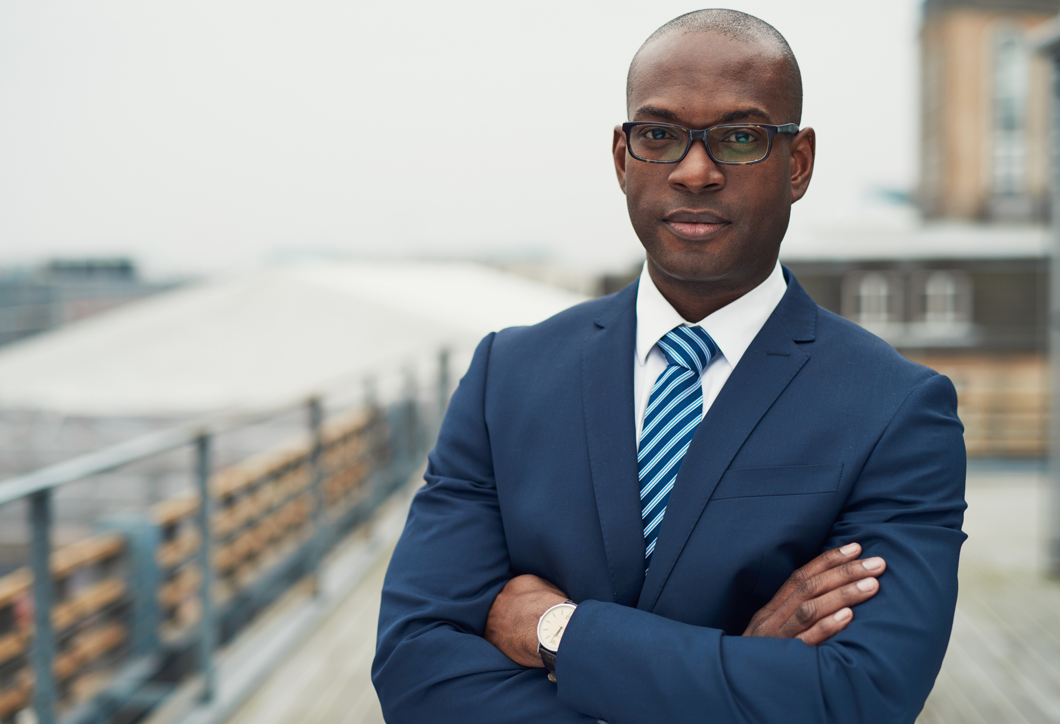 A man wearing a suit and tie stands with arms crossed on the roof of a building.