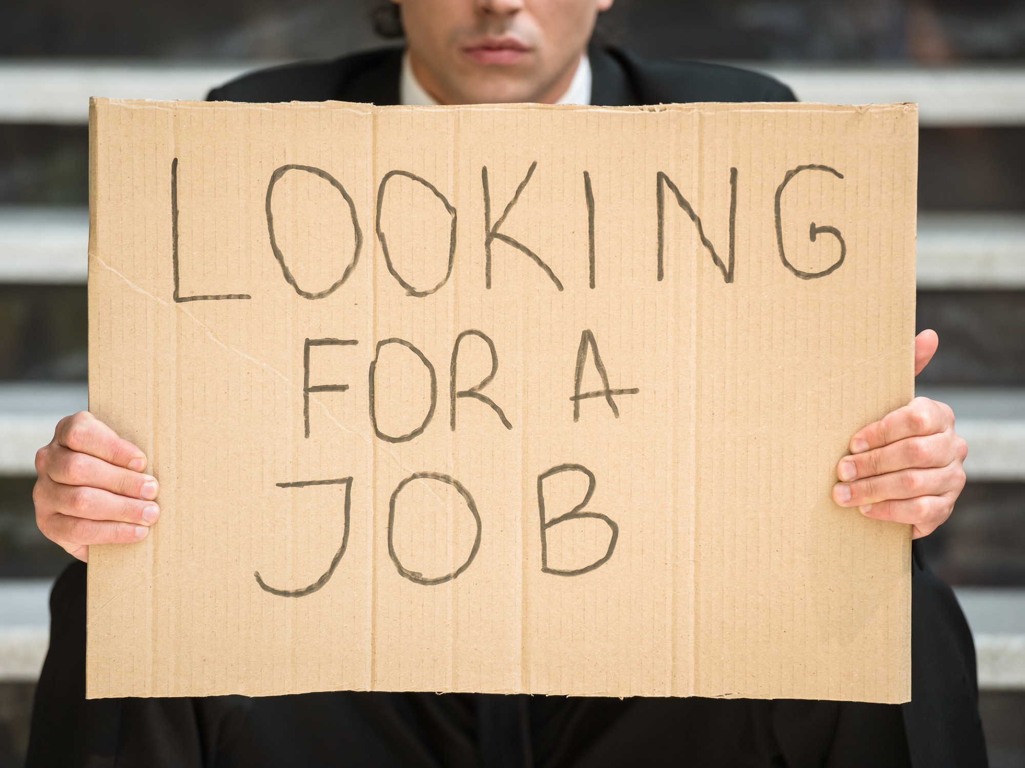 A businessman in a suit holding up a cardboard sign that reads, Looking for a job.