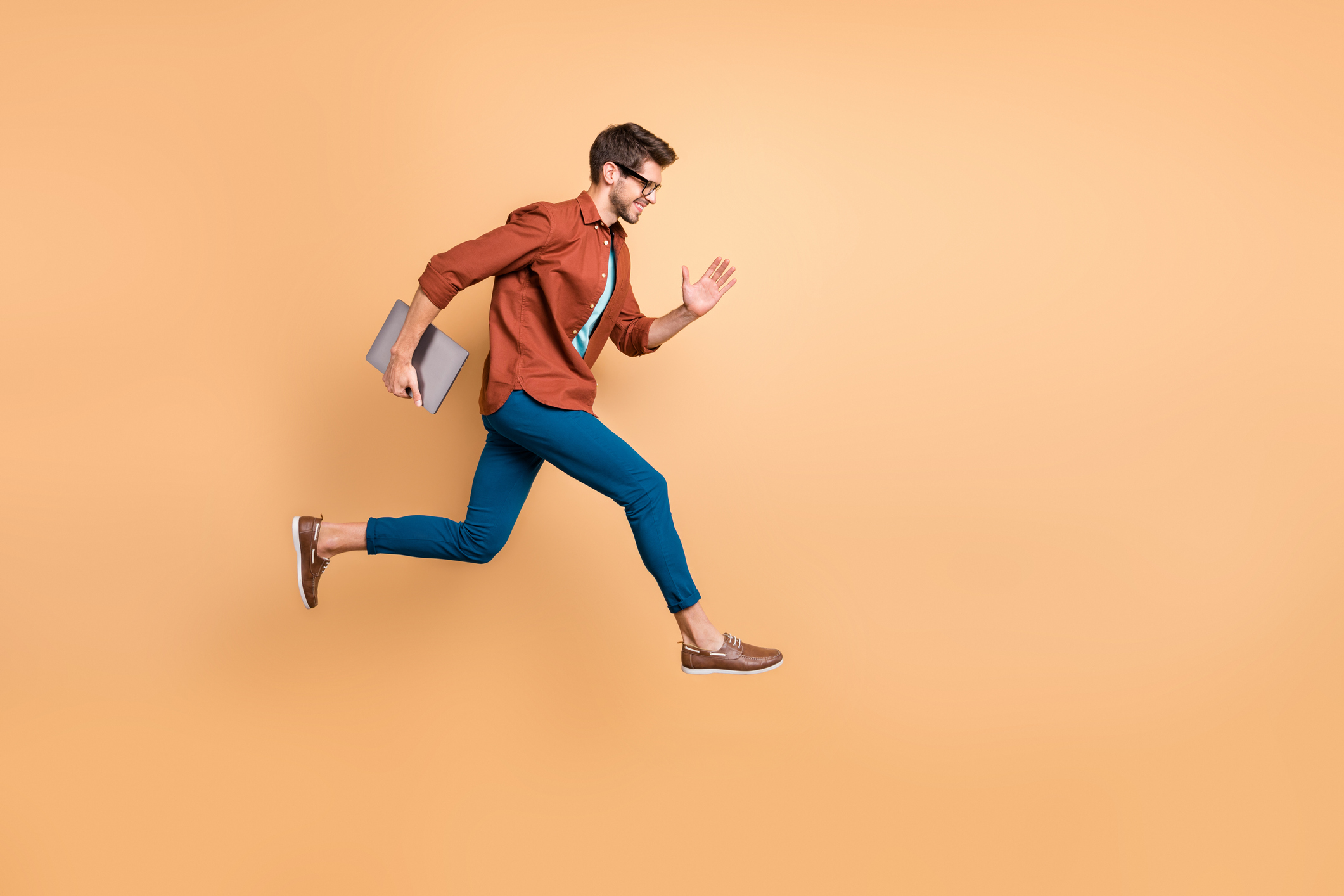A young man carries a laptop and runs  in mid air against a beige background.