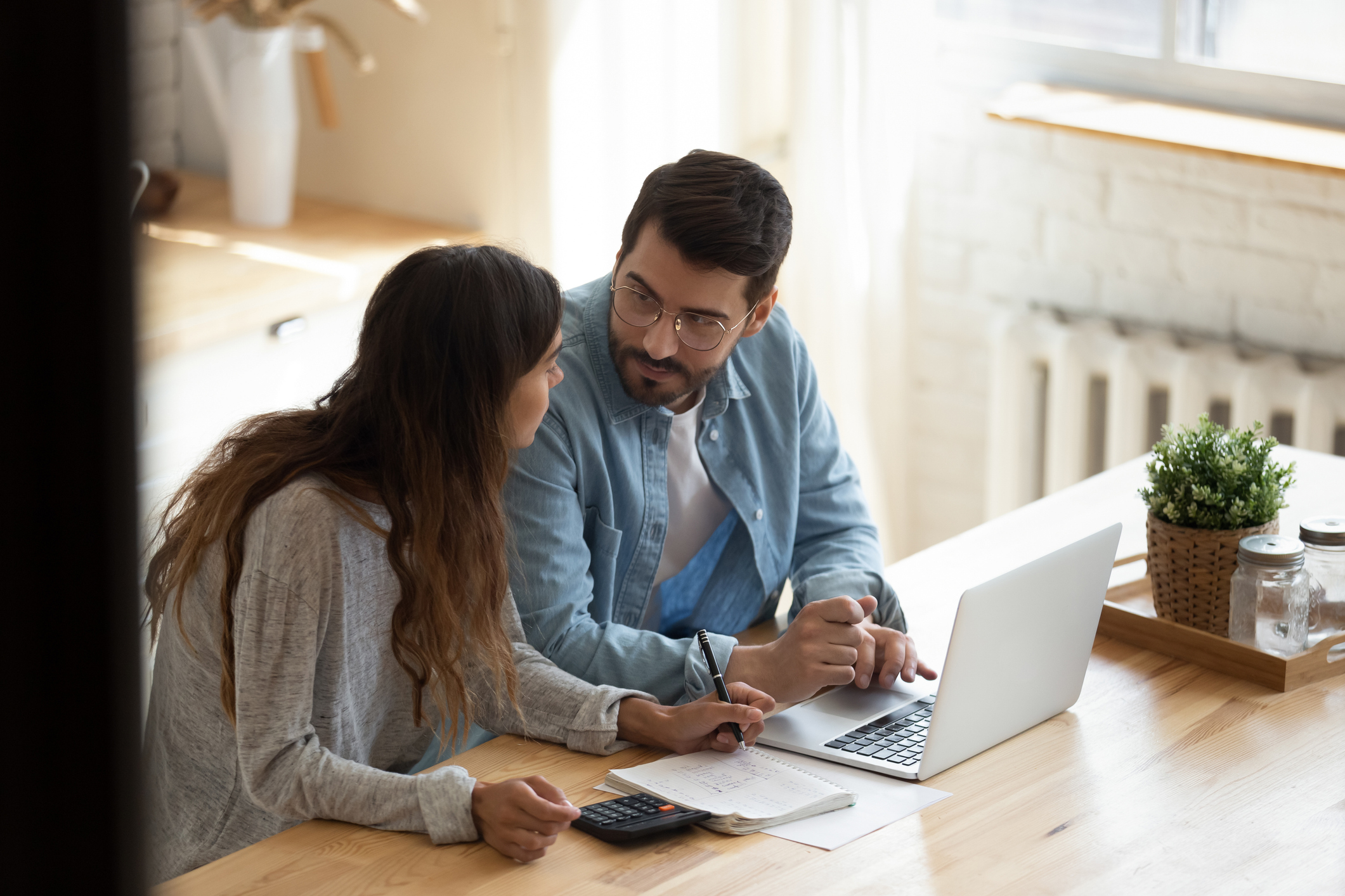 Man at laptop sitting next to woman taking notes in notebook next to calculator
