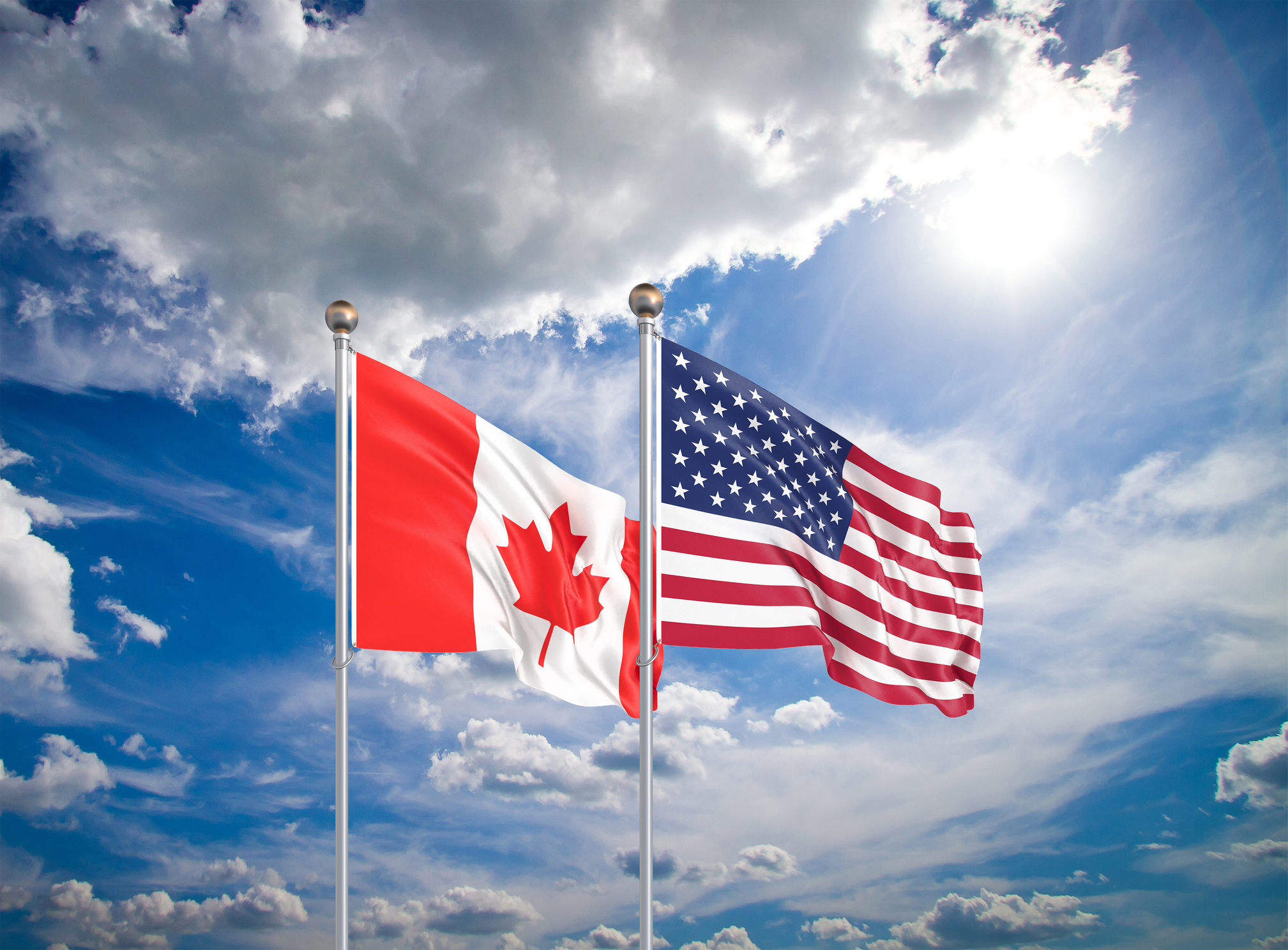 US and Canadian flags fly side by side against a cloudy blue sky.