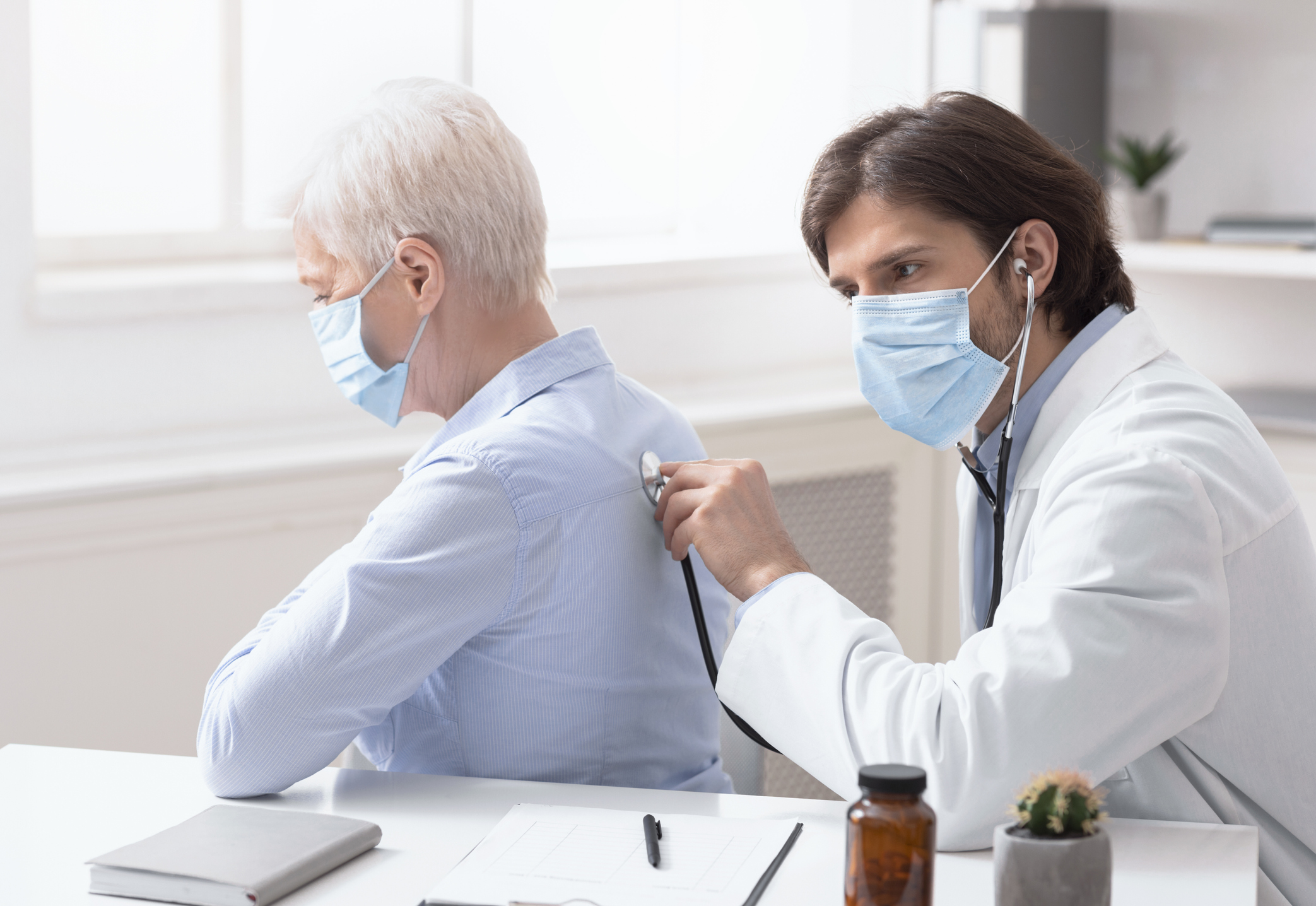 Masked doctor pressing stethoscope into masked older patient's back
