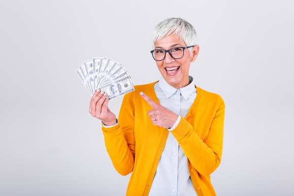 A happy senior woman is smiling and pointing to a handful of cash.