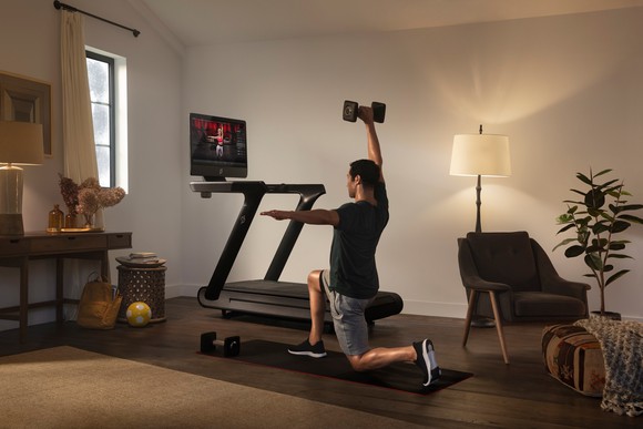 Person at home lifting hand weight next to treadmill with video screen on.