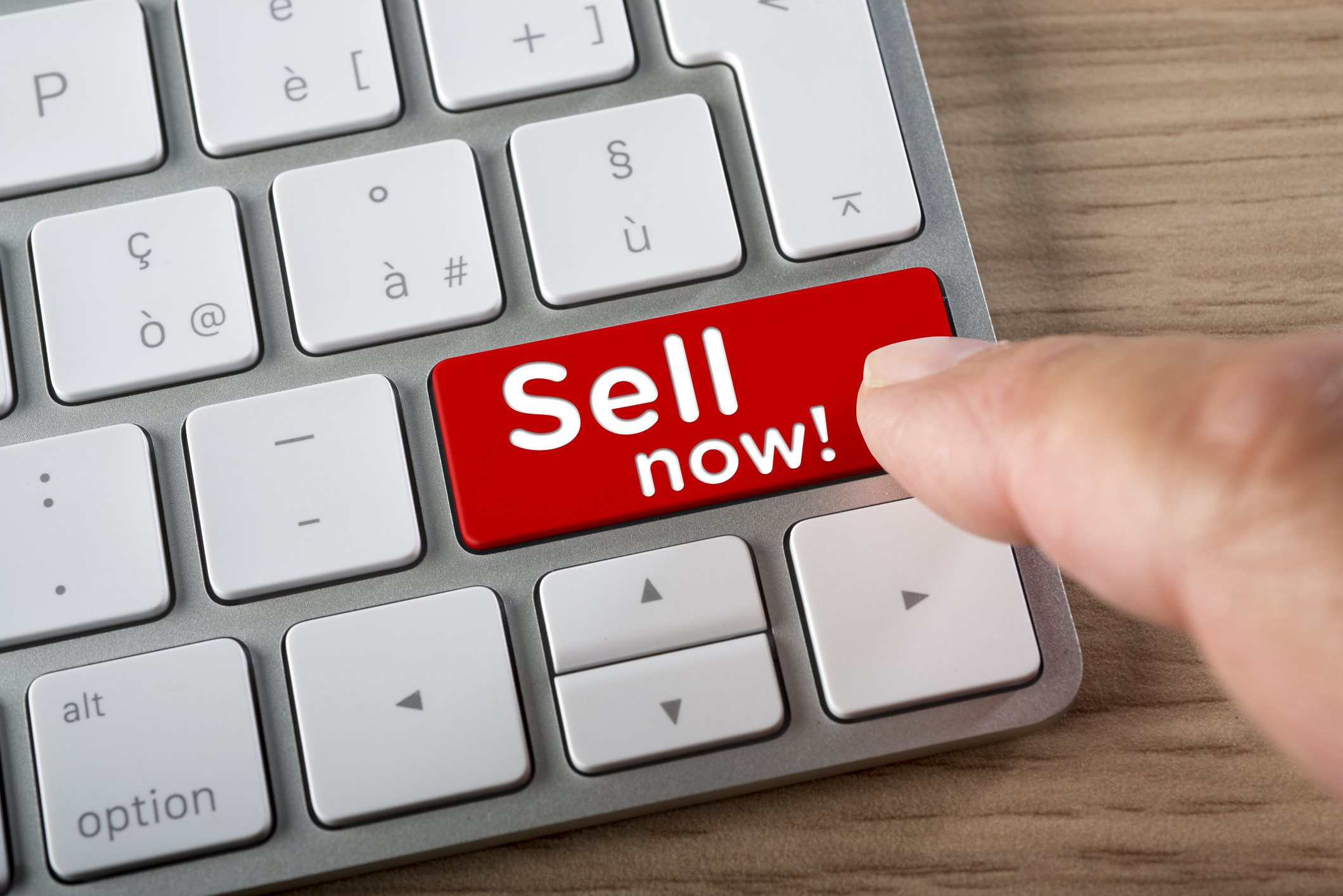 A person pressing a red key on a computer keyboard that emphatically reads, Sell now!