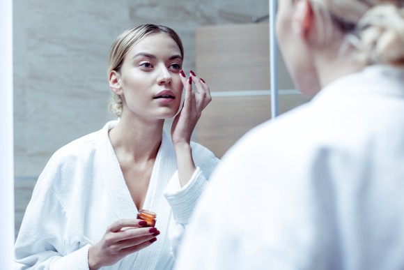 A woman in a robe applying an eye cream while looking in a mirror