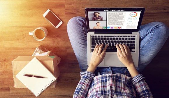 A person sitting cross-legged working remotely with a laptop showing video conferencing and workplace collaboration software.