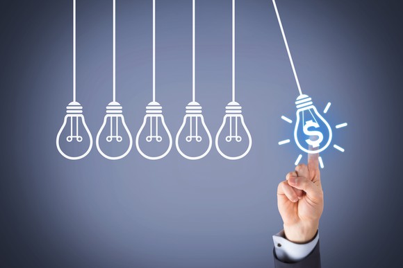 Finger pointing to drawing of a pendulum light bulb with a dollar sign in it next to a row of ordinary pendulum bulbs. 