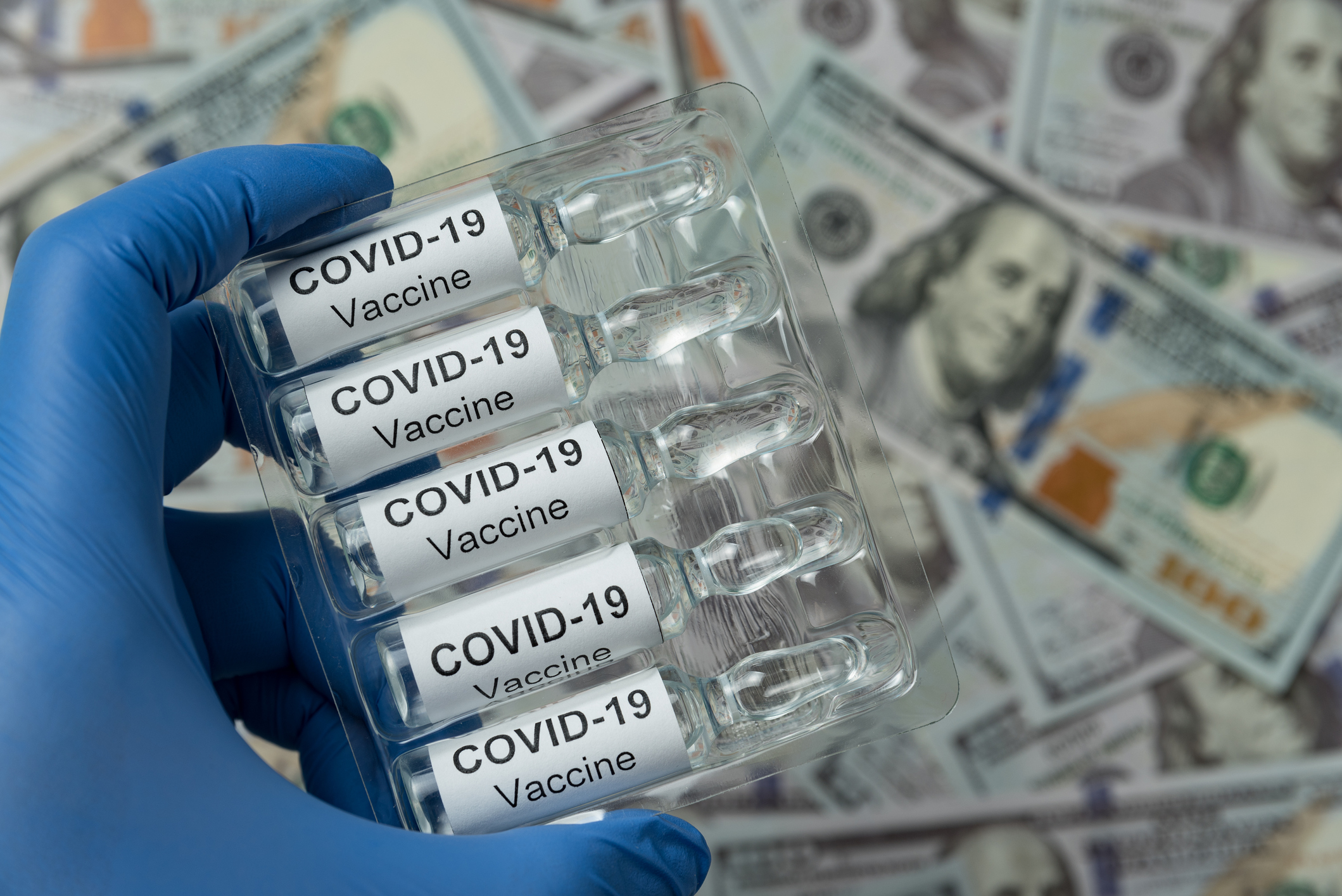 Gloved hand holding vials labeled COVID-19 vaccine over a pile of hundred-dollar bills