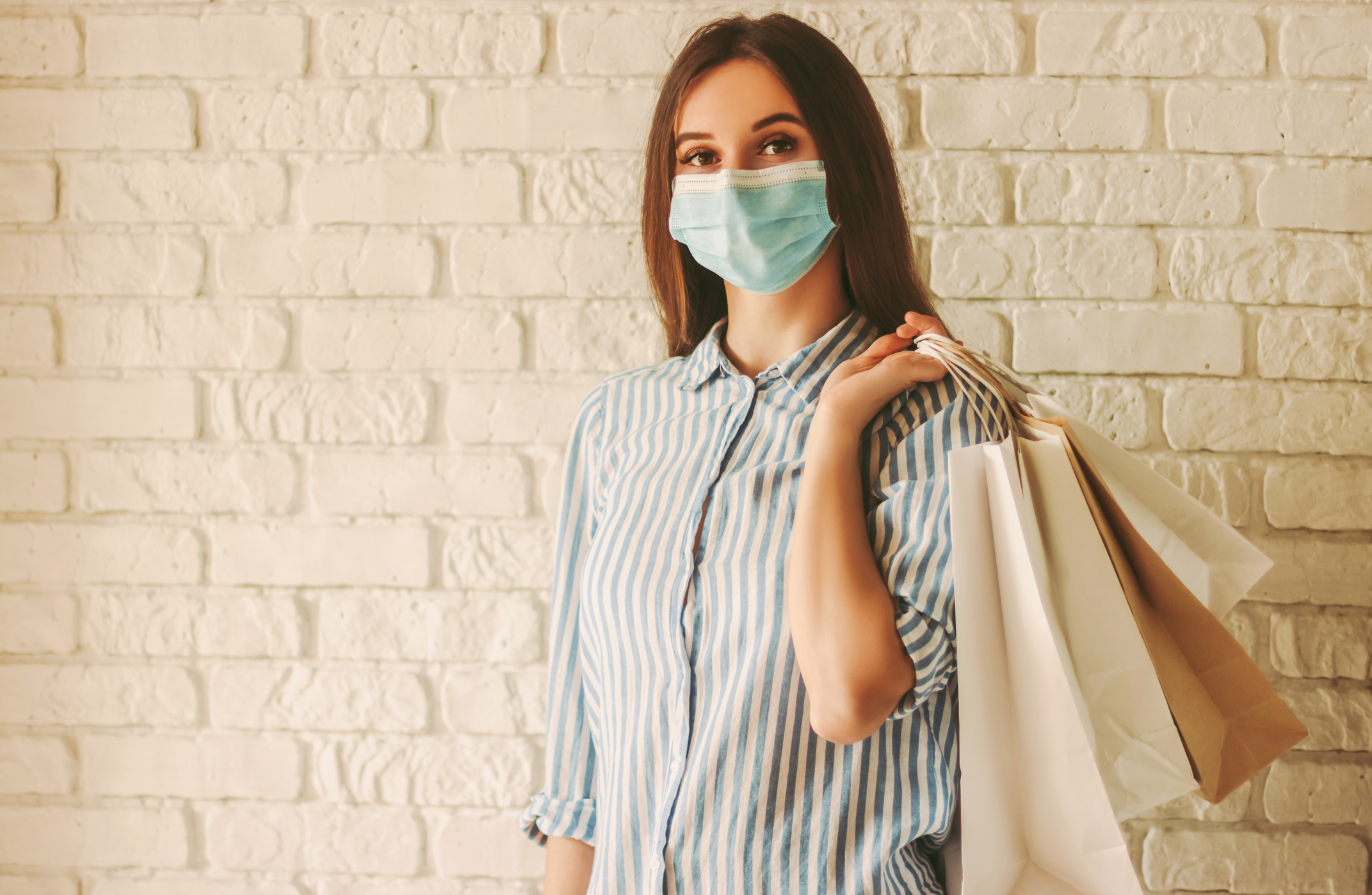 A woman, wearing a mask, holds shopping bags as she stands in front of a brick wall.