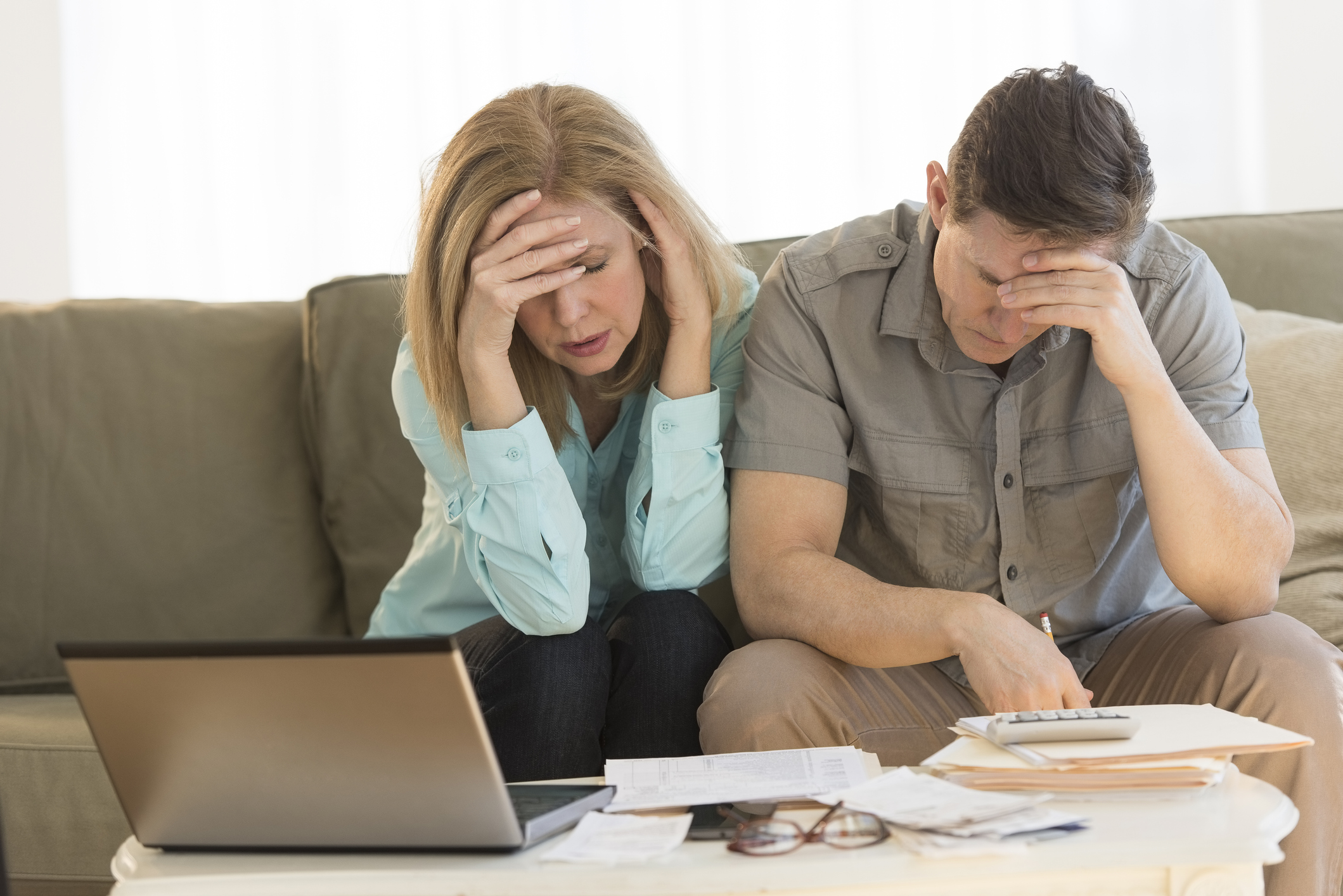 Older man and woman holding their heads while sitting on a couch; in front of them is a table with a laptop, folders, papers, eyeglasses, and calculator