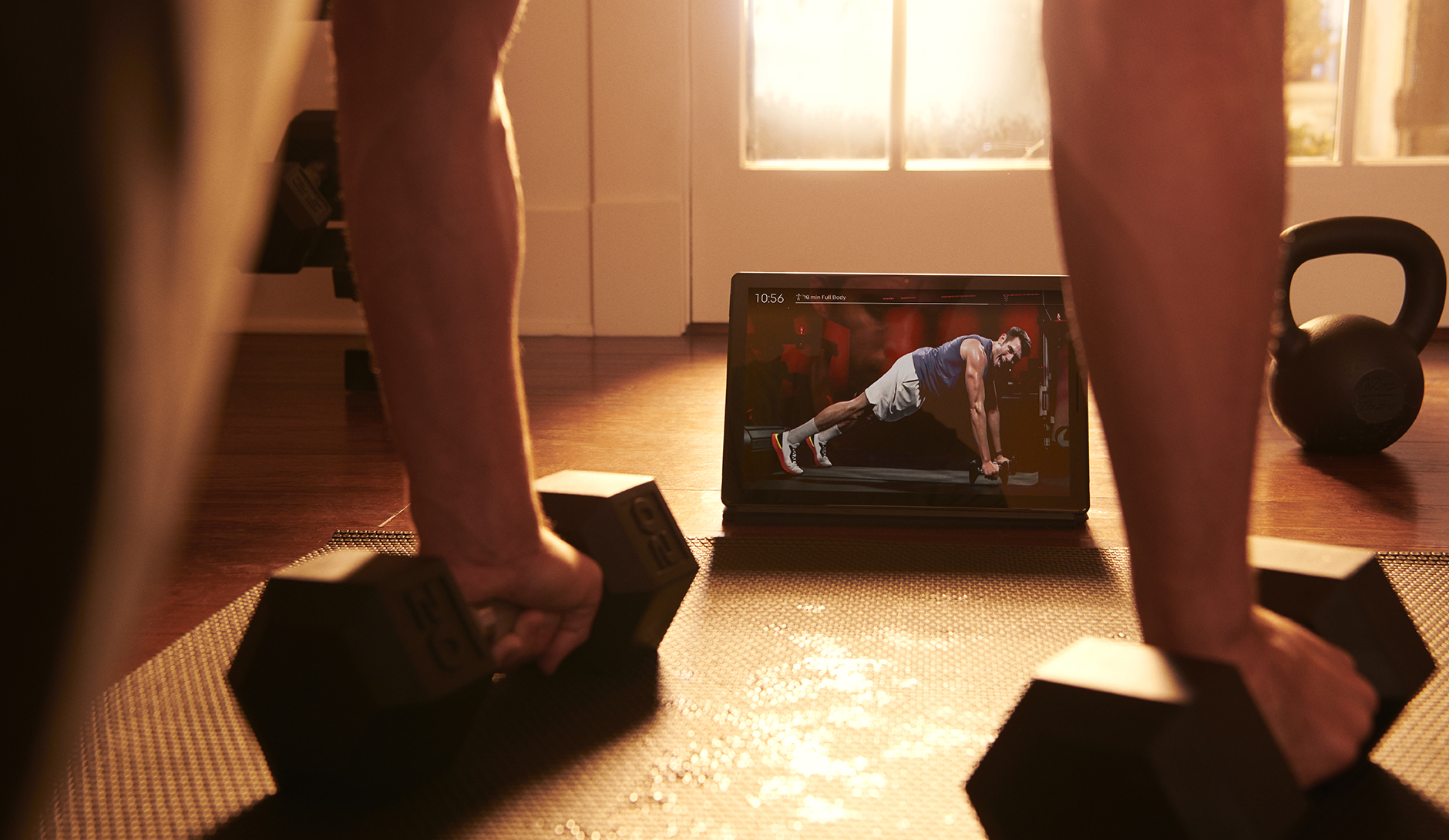 A person does an exercise with two barbells while watching Peloton video content.