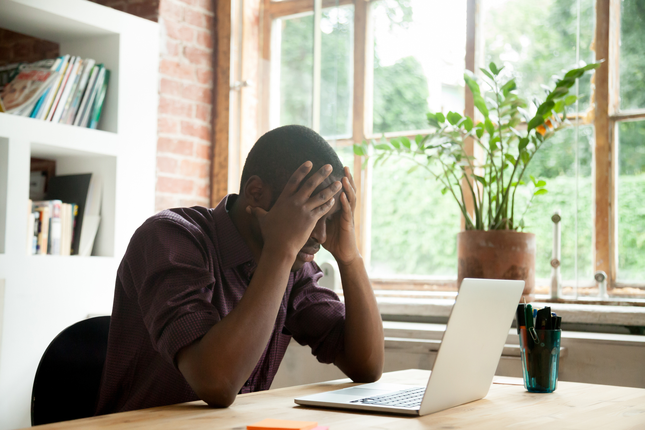 Person seated at a table in front of a laptop, holding their head in their hands.