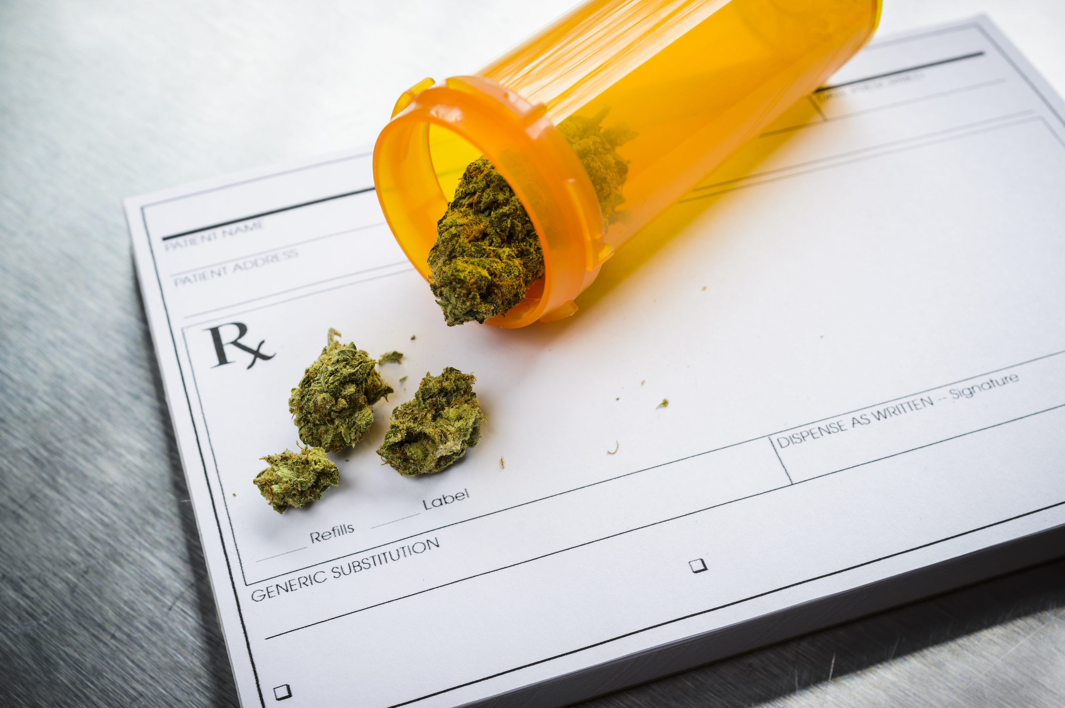 A tipped over prescription bottle containing dried cannabis buds lying atop a doctors prescription pad.