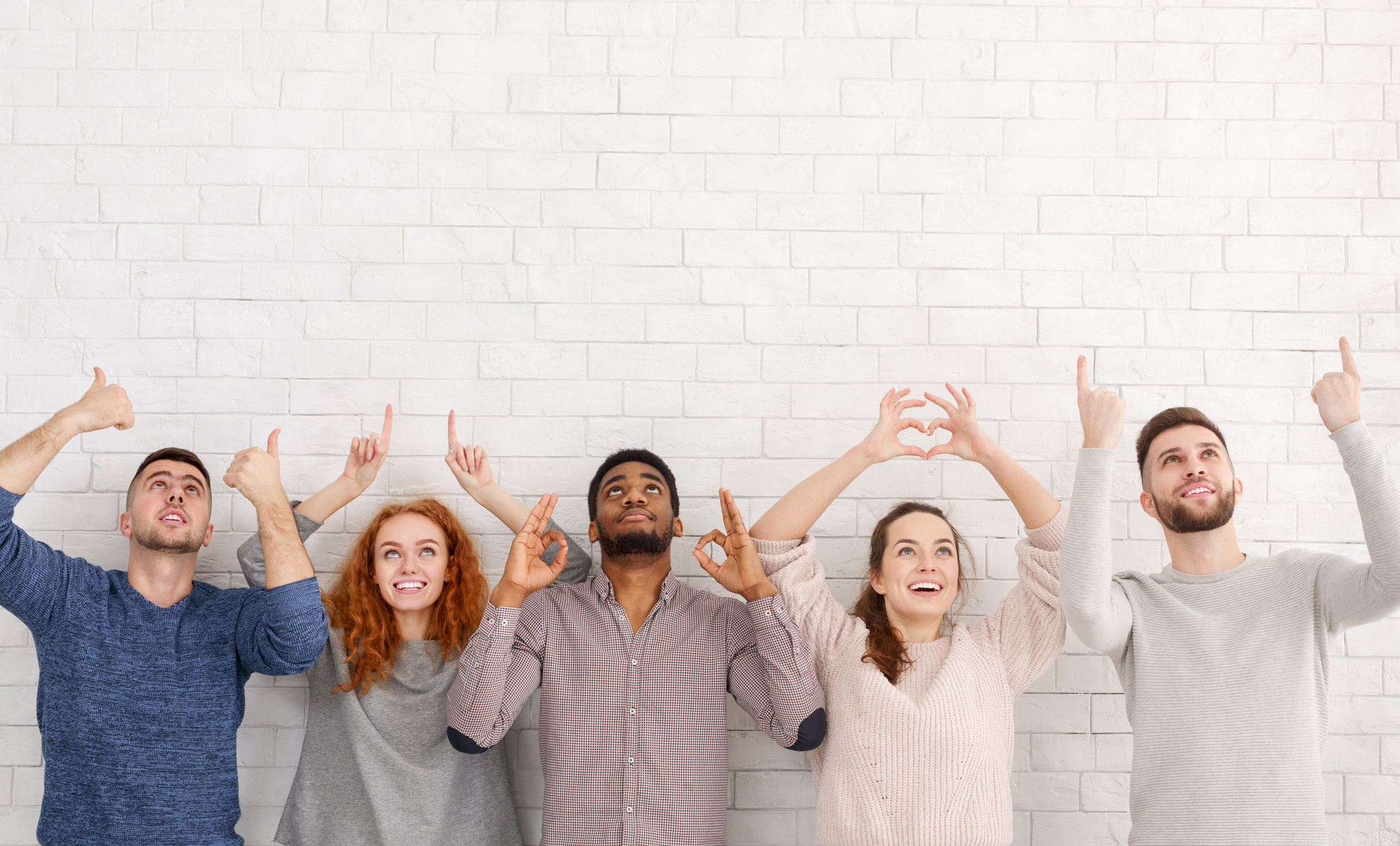 A group of young millennials look upwards against a white wall making hand gestures.