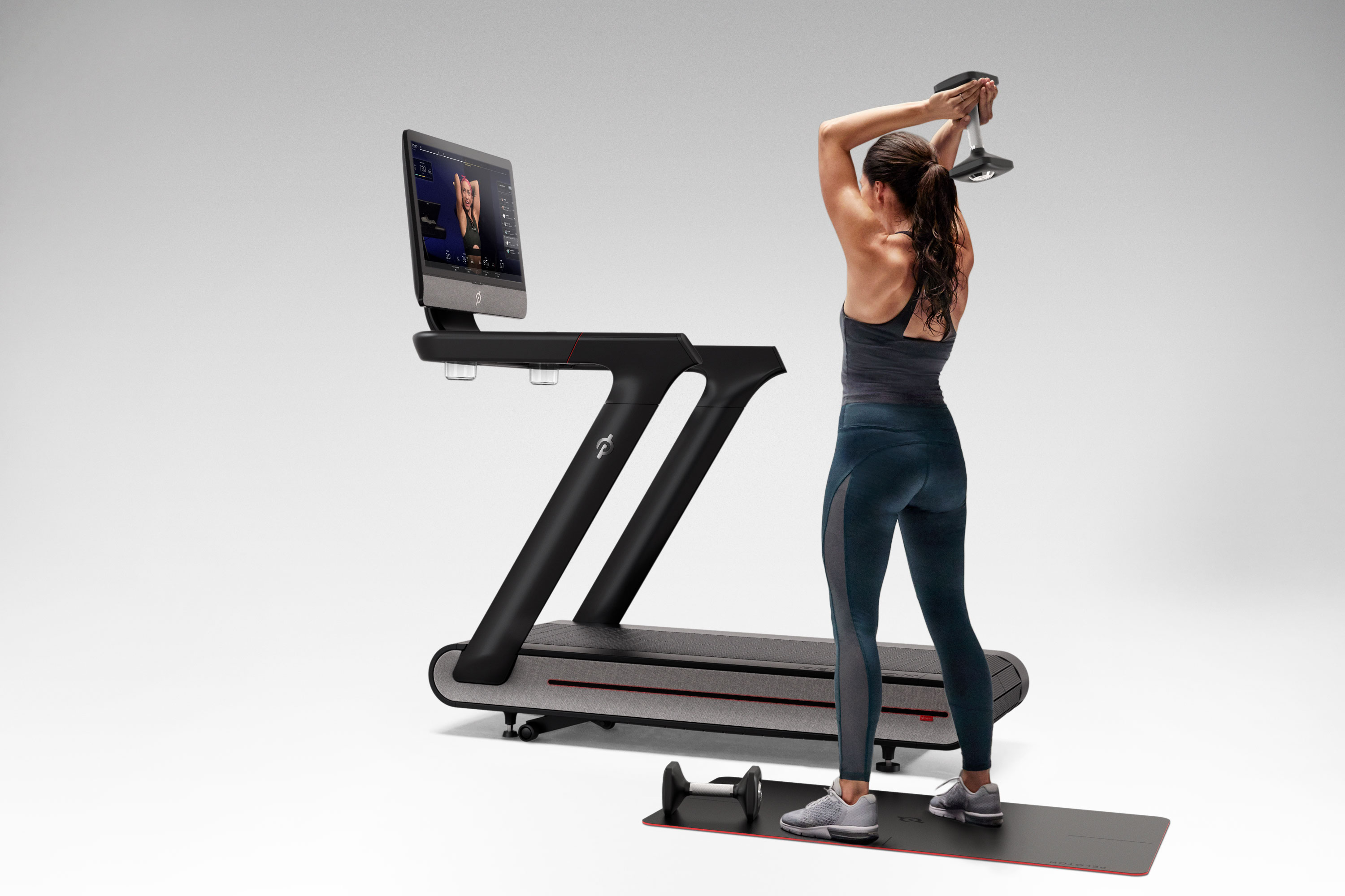 A woman lifting weights next to a Peloton Tread product.