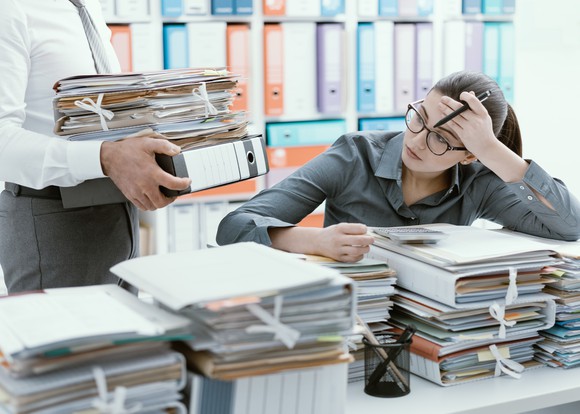 A frustrated woman with a desk full of paperwork and a coworker bringing more
