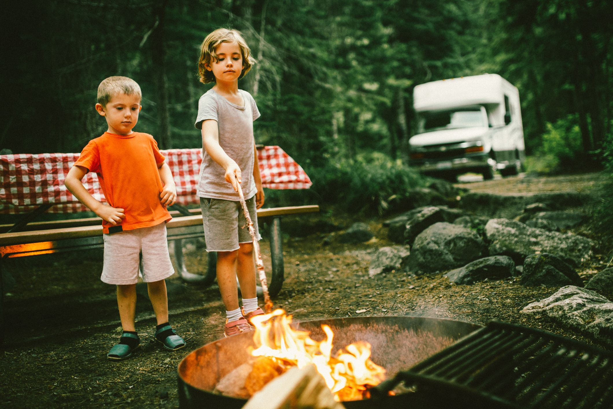 Two kids at a campfire, with an RV in the background.