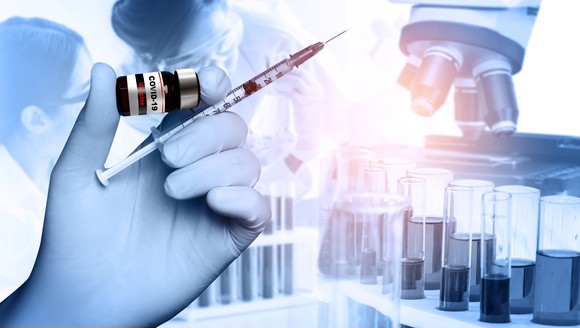 Gloved hand holding a vaccine vial and syringe with scientists, a microscope, and test tubes in the background.