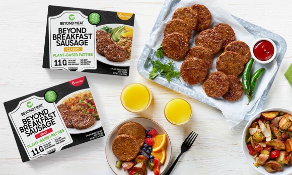 Beyond Breakfast Sausage patties, packaging, and two glasses of orange juice on a white tabletop.