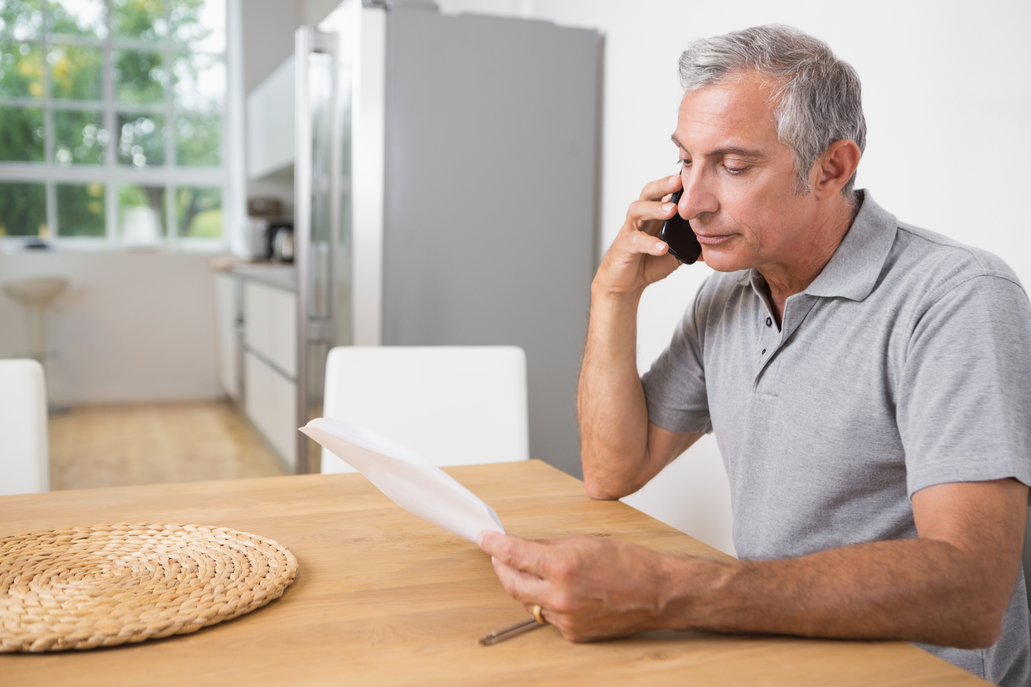 An older person sitting at a table holding a document with one hand and a phone to their ear with the other.