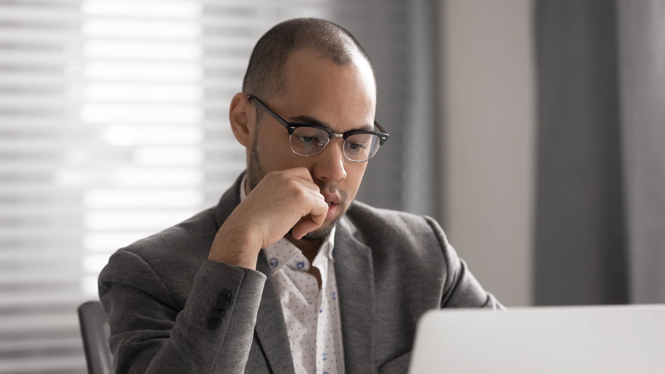 A young adult male in a business casual outfit pores over something on his laptop.
