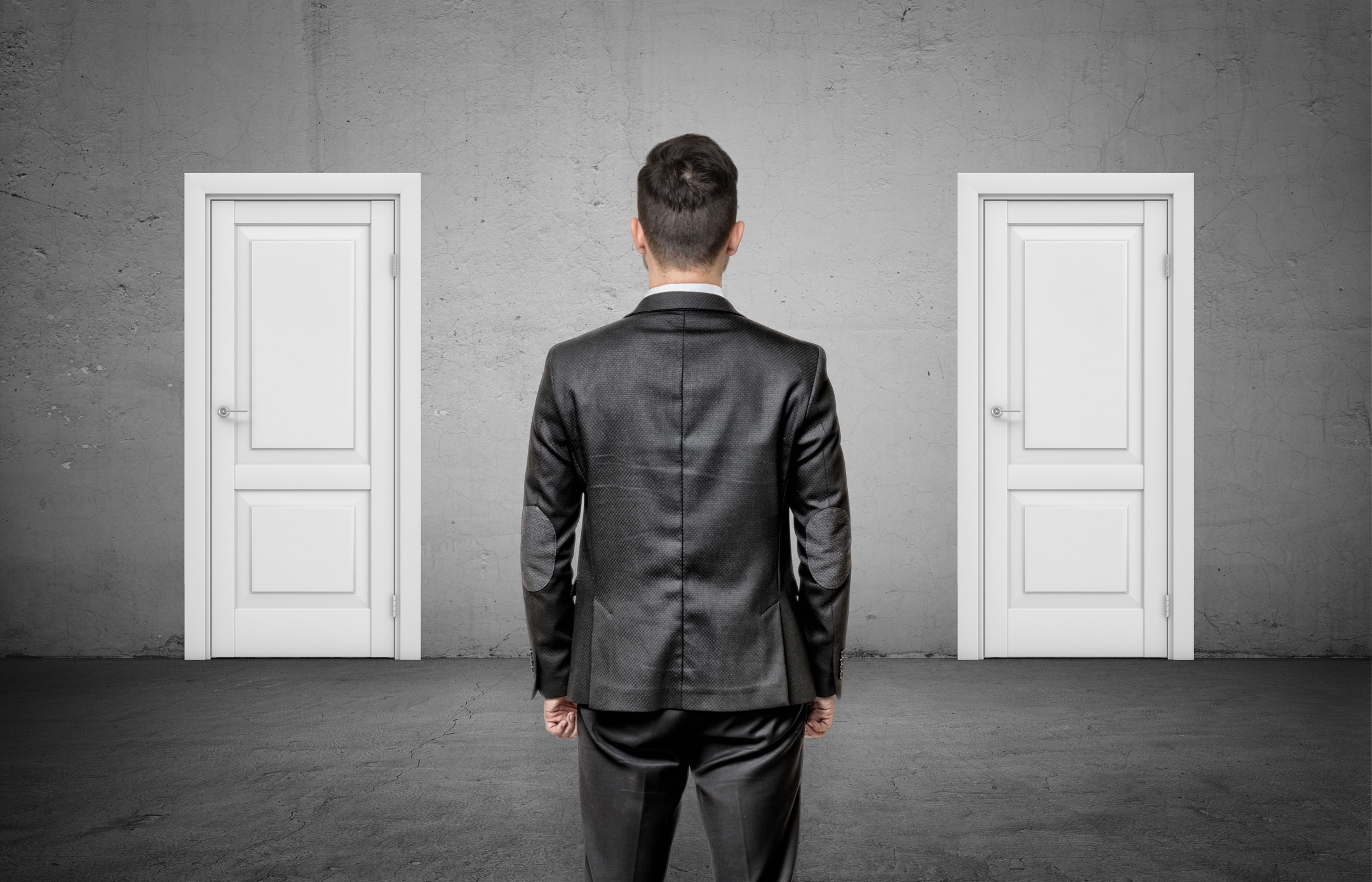 A young man in a suit seen from behind looking at two identical white doors. 