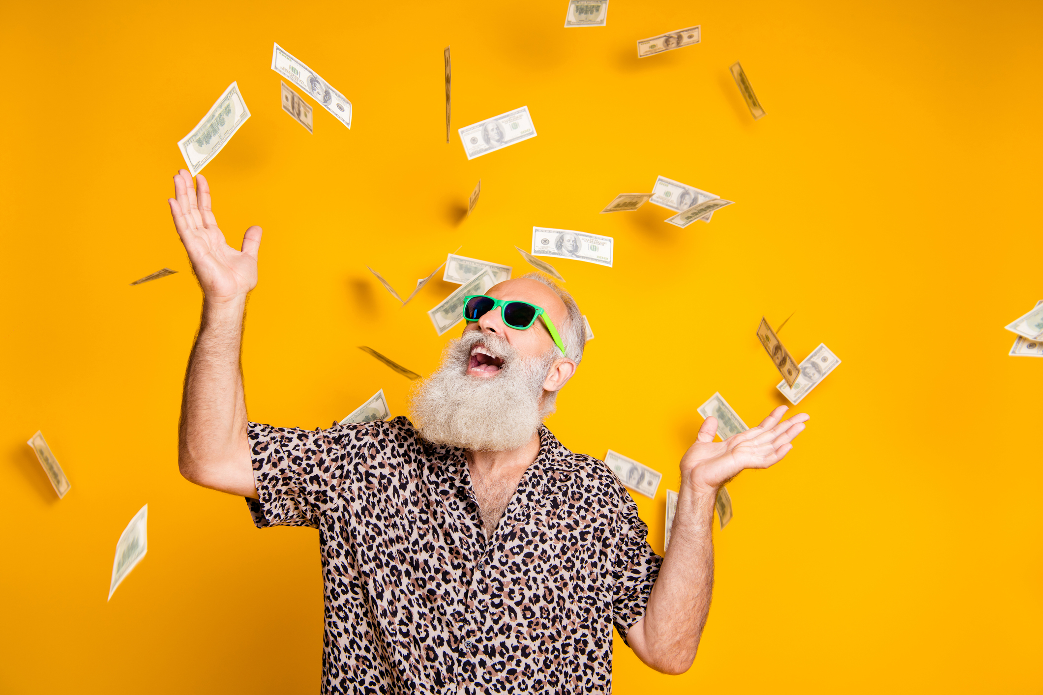 An elderly, bearded man with sunglasses stands under raining cash bills against an orange background.