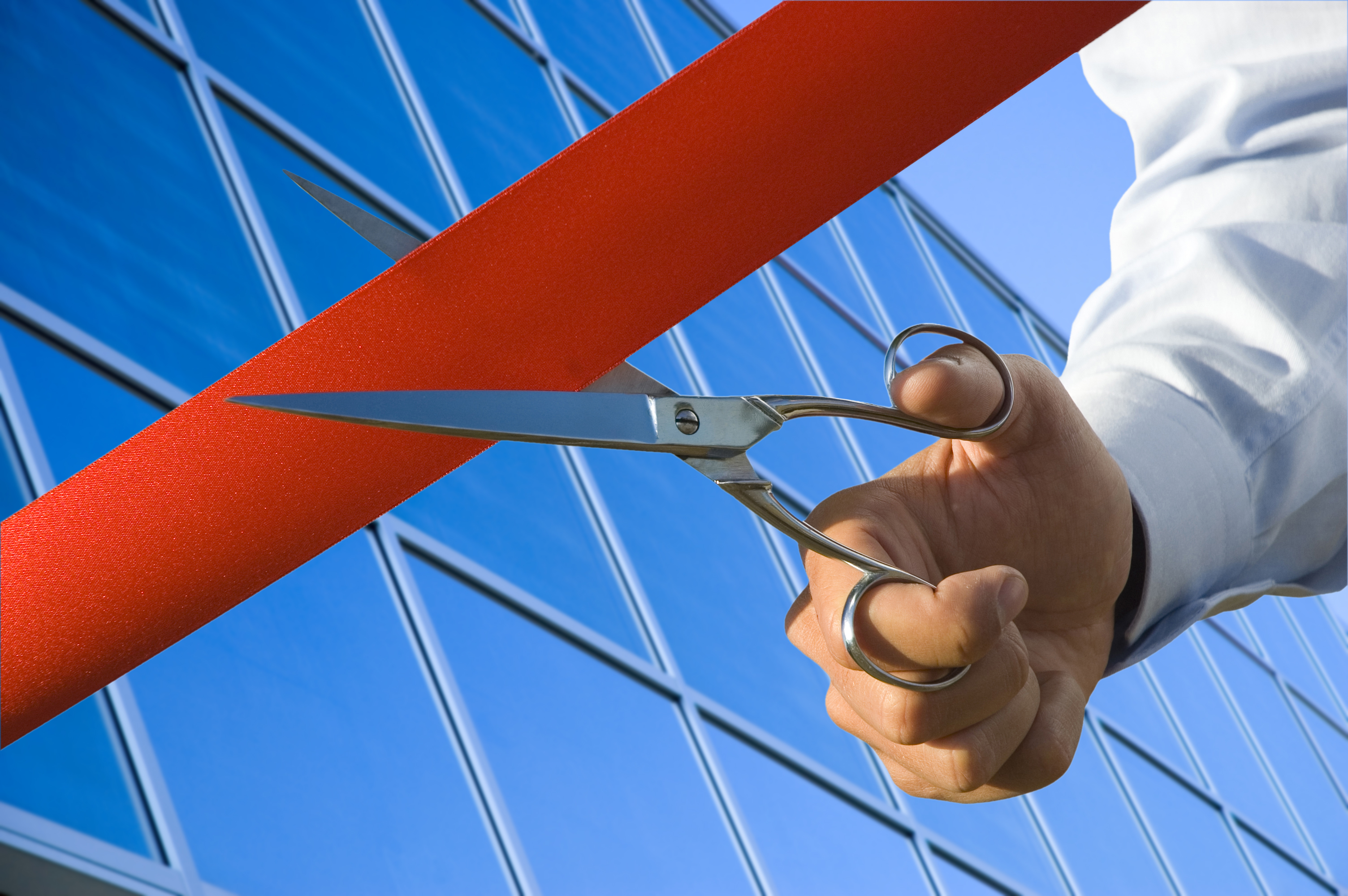 A man's hand holding a pair of scissors cutting a red ribbon outside an opening business location.