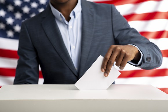 A person putting ballot in box with a U.S. flag in the background.