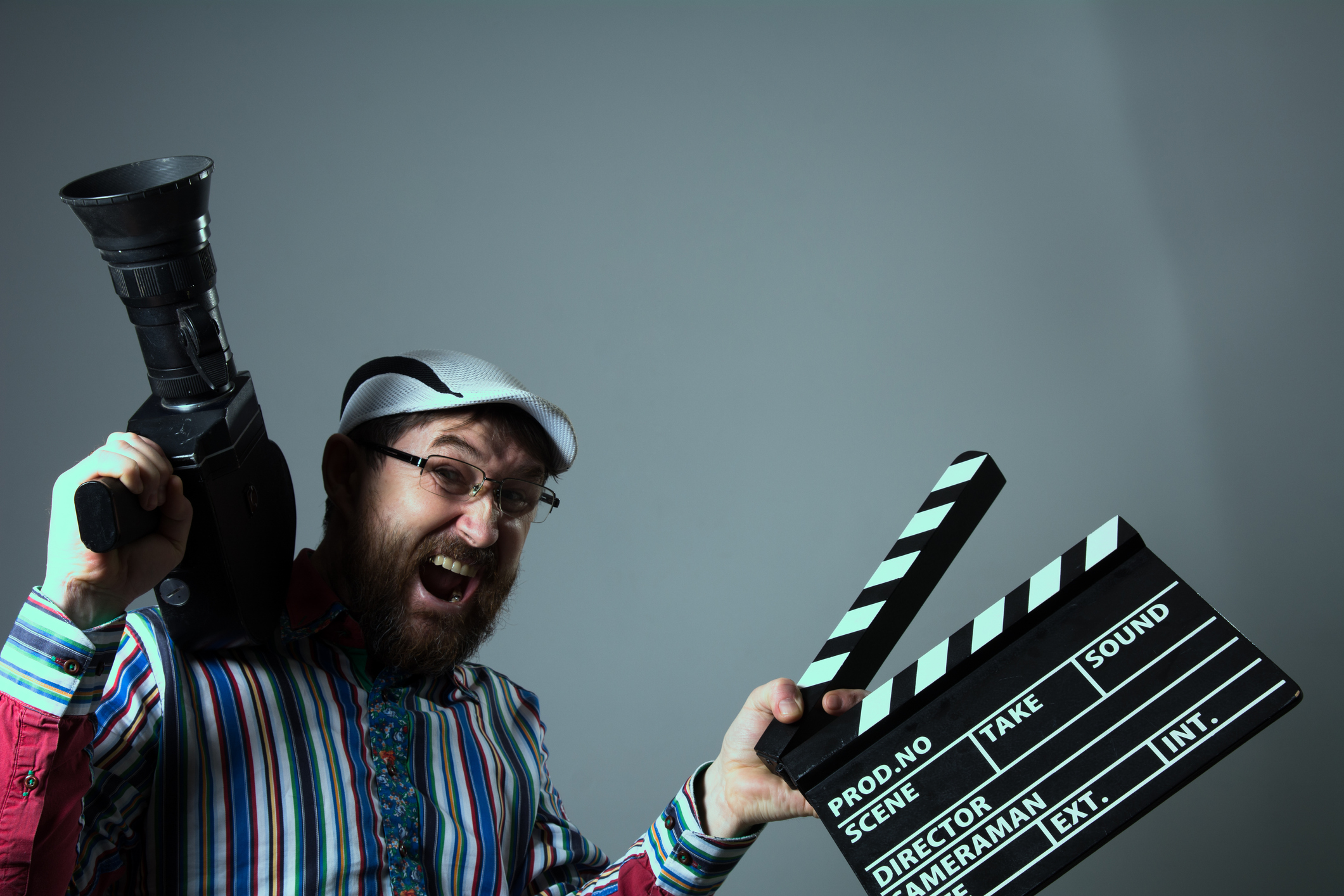 A beared man smiles at the camera, holding a movie camera in one hand and a clapper board in the other.