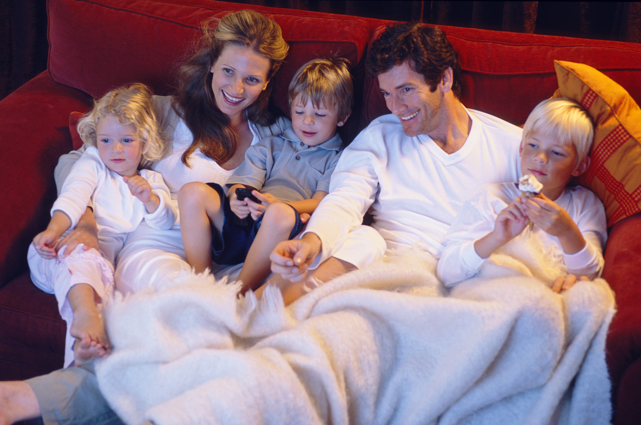 A husband and wife and their three young children sitting on the couch under a blanket watching television.