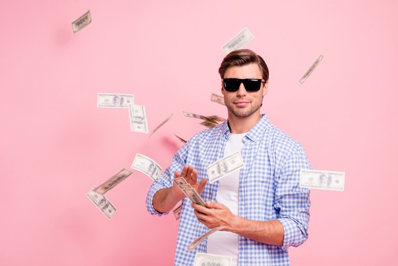 A man standing in front of a pink background and wearing sunglasses throwing money into the air