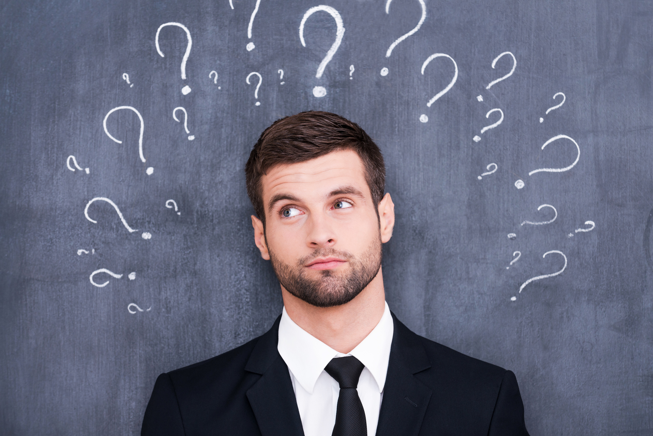 Man standing in front of a chalkboard with question marks drawn on the board around his head. 